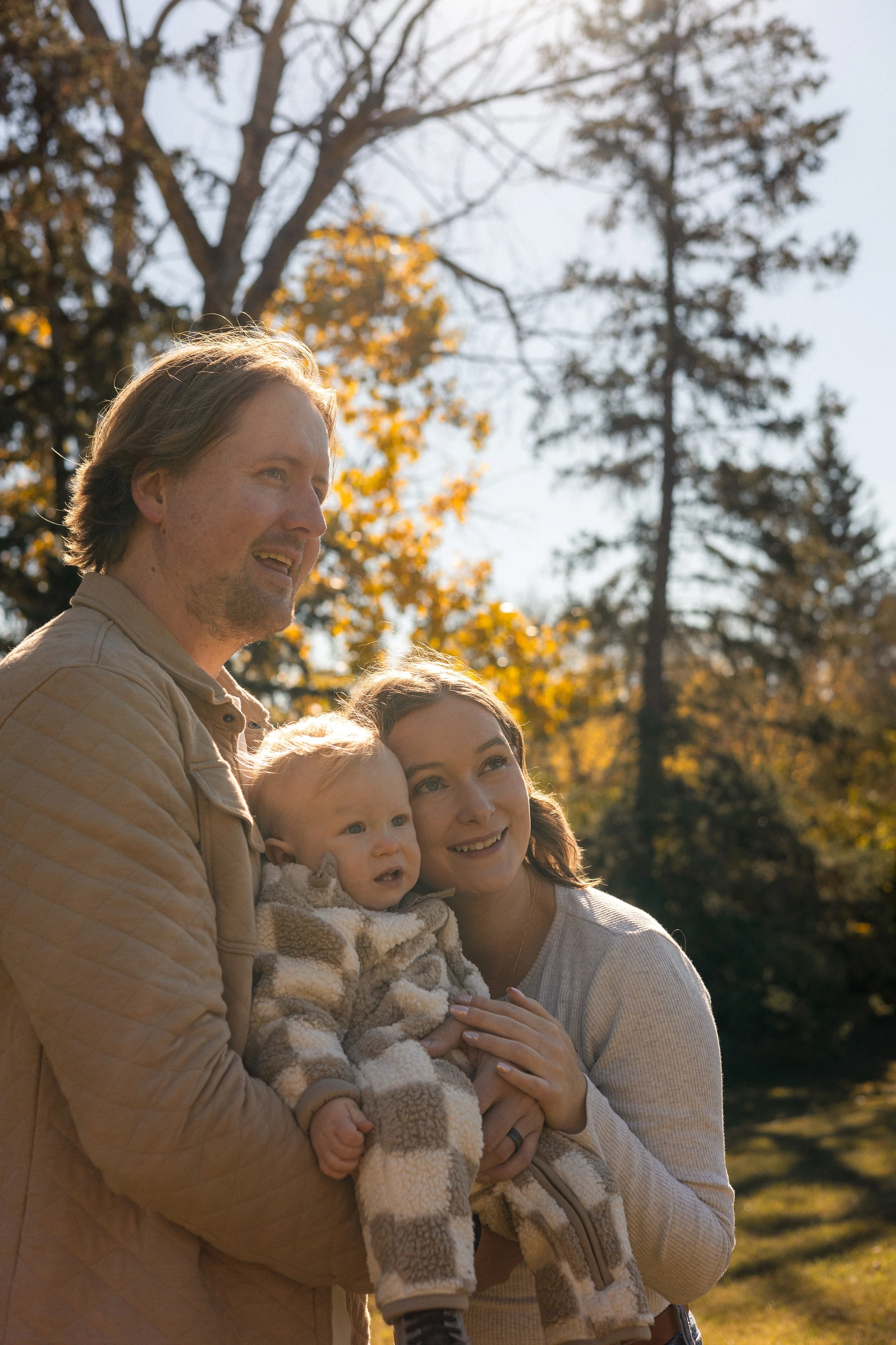 A family of four standing outdoors in a park during autumn, with trees showing fall foliage in the background. The man, woman, and two children are smiling and happy.