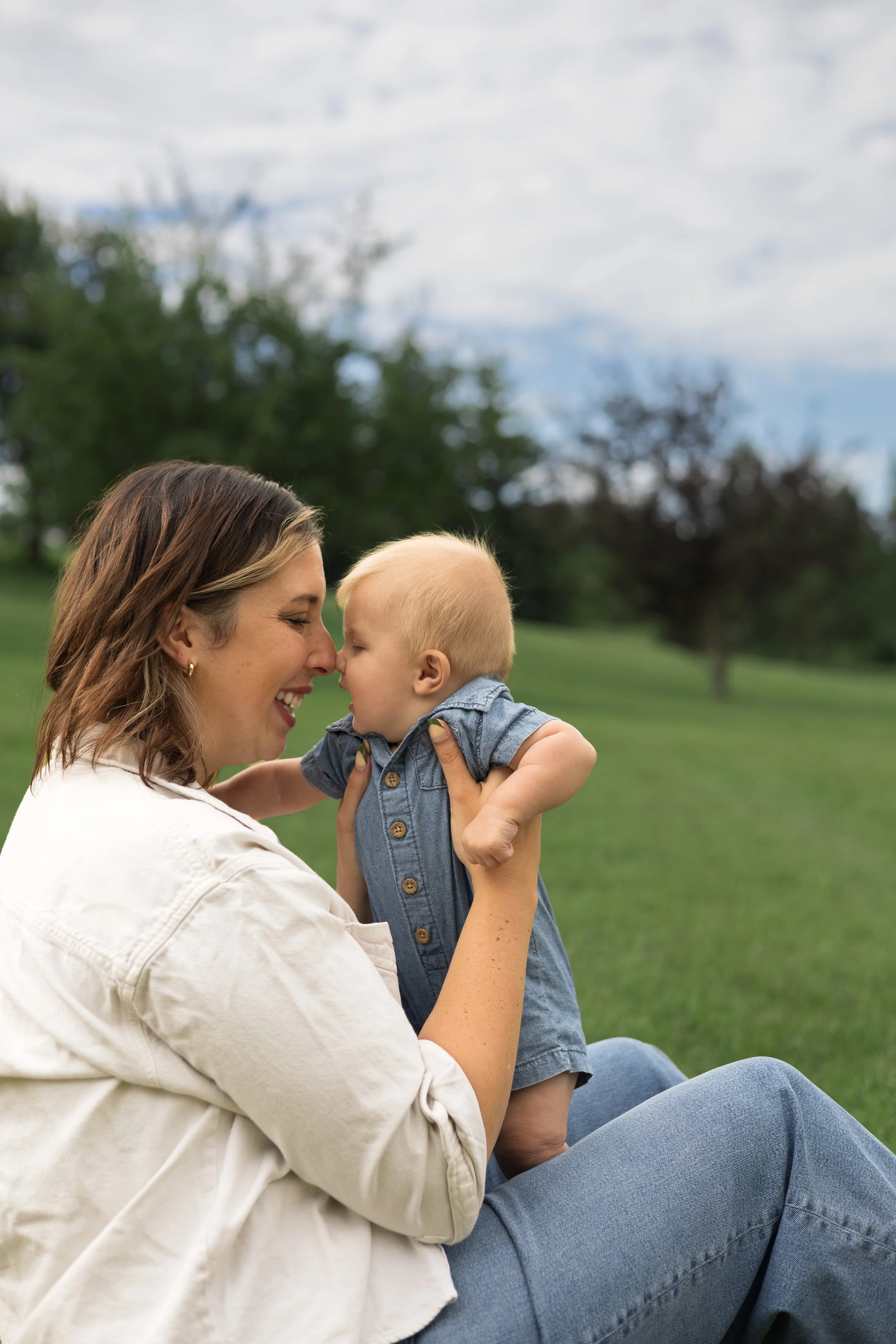 A woman with brown hair and wearing a white jacket is holding a young child with blonde hair in a park. They are touching foreheads and smiling at each other, with trees and grass in the background.  Location is Confederation Park in Calgary, Alberta