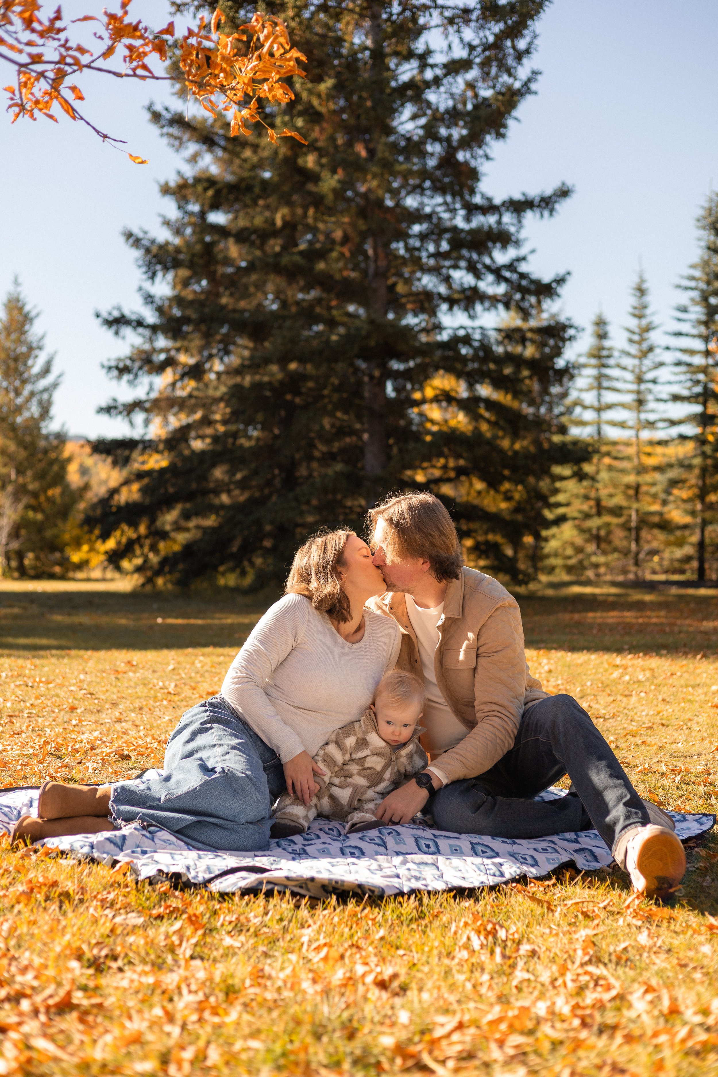 A family of three sitting on a blanket on fallen autumn leaves in a park, sharing a kiss with a tree in the background.