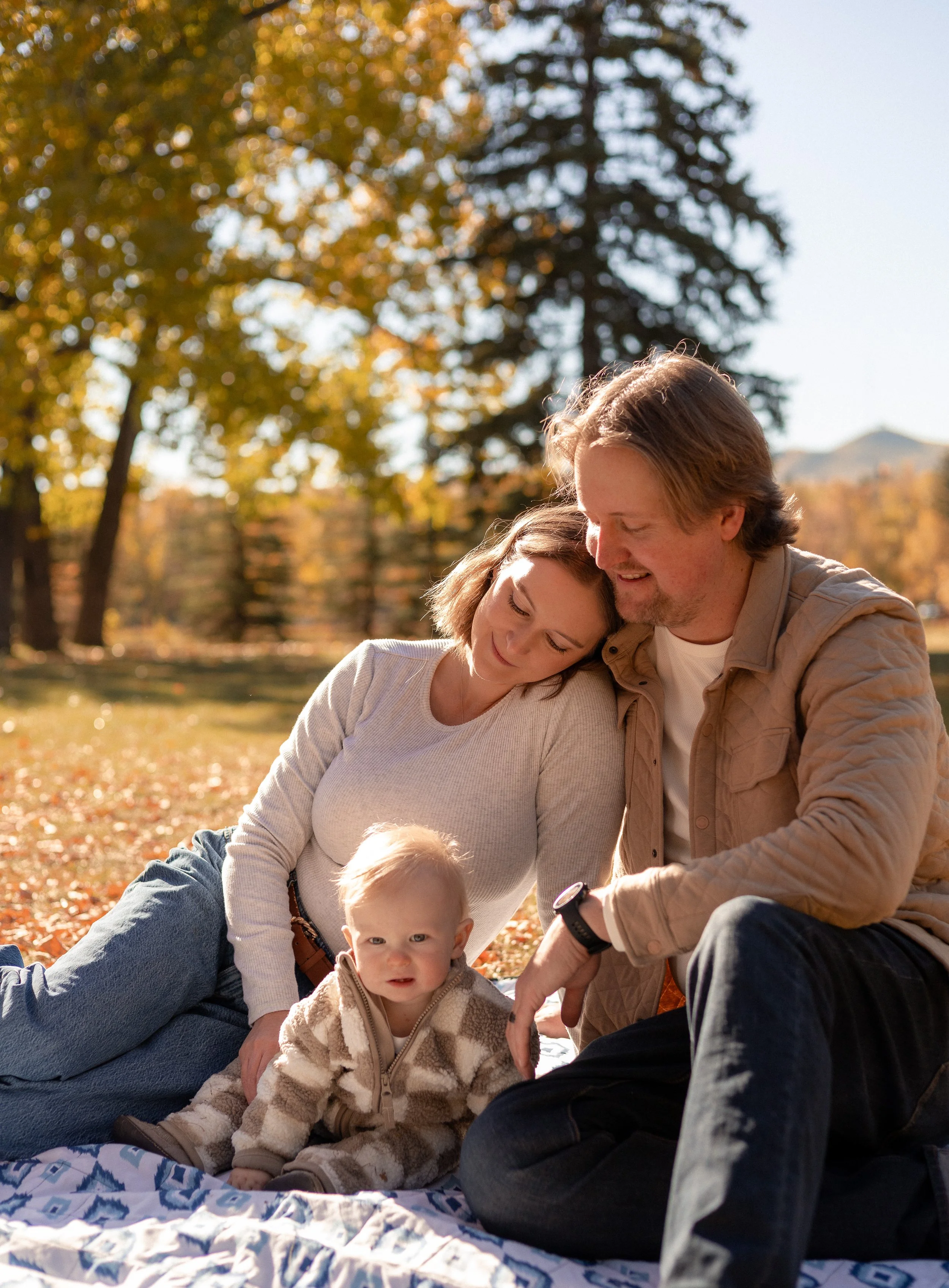 A family of three, including a baby, enjoying an outdoor fall day. The mother and father are sitting close together on a blanket, with the mother resting her head on the father's shoulder. They are surrounded by trees with autumn leaves, and a mounta
