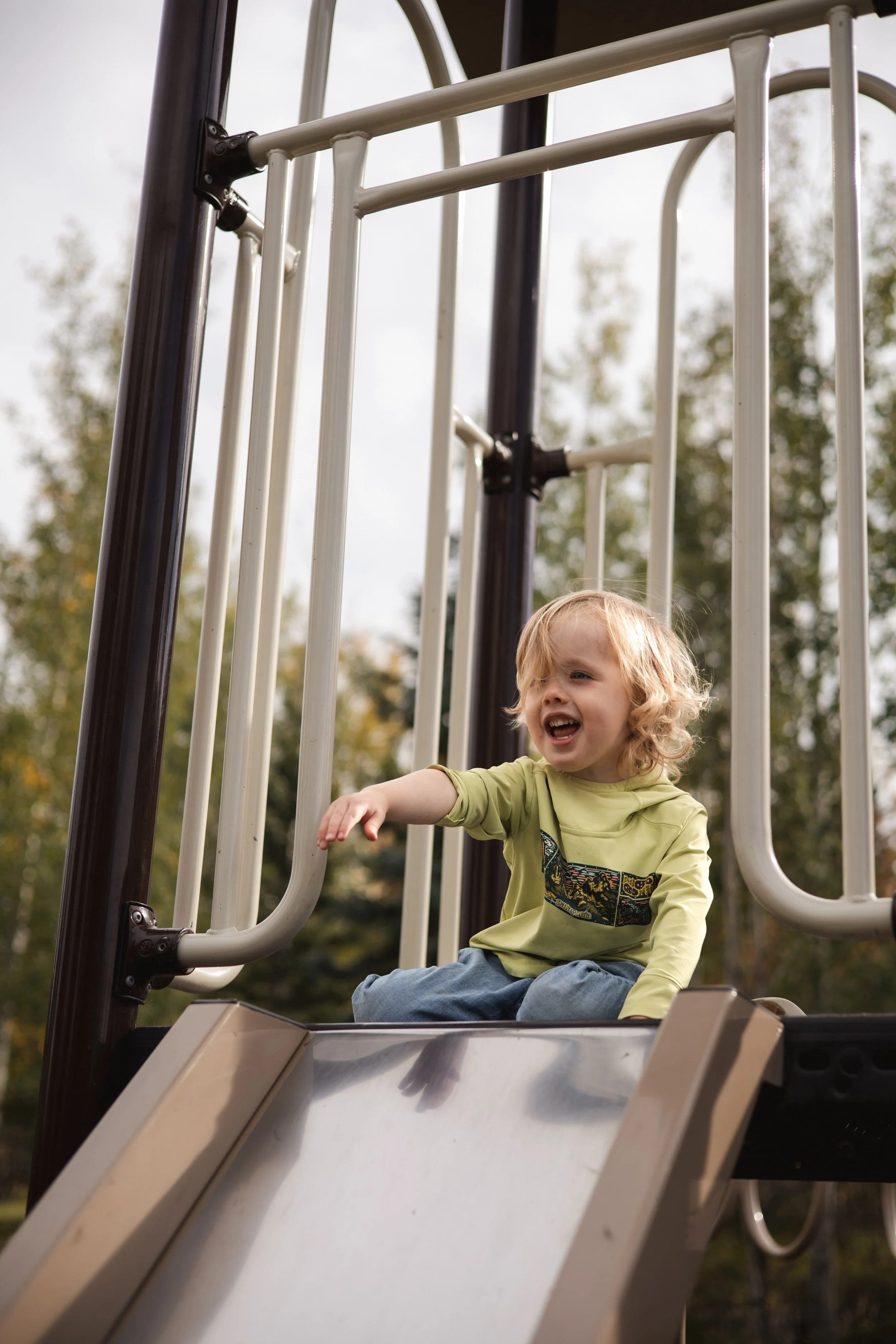 A young child with blonde hair and a yellow hoodie sitting at the top of a playground slide, smiling and pointing with one hand, outdoors with trees in the background.