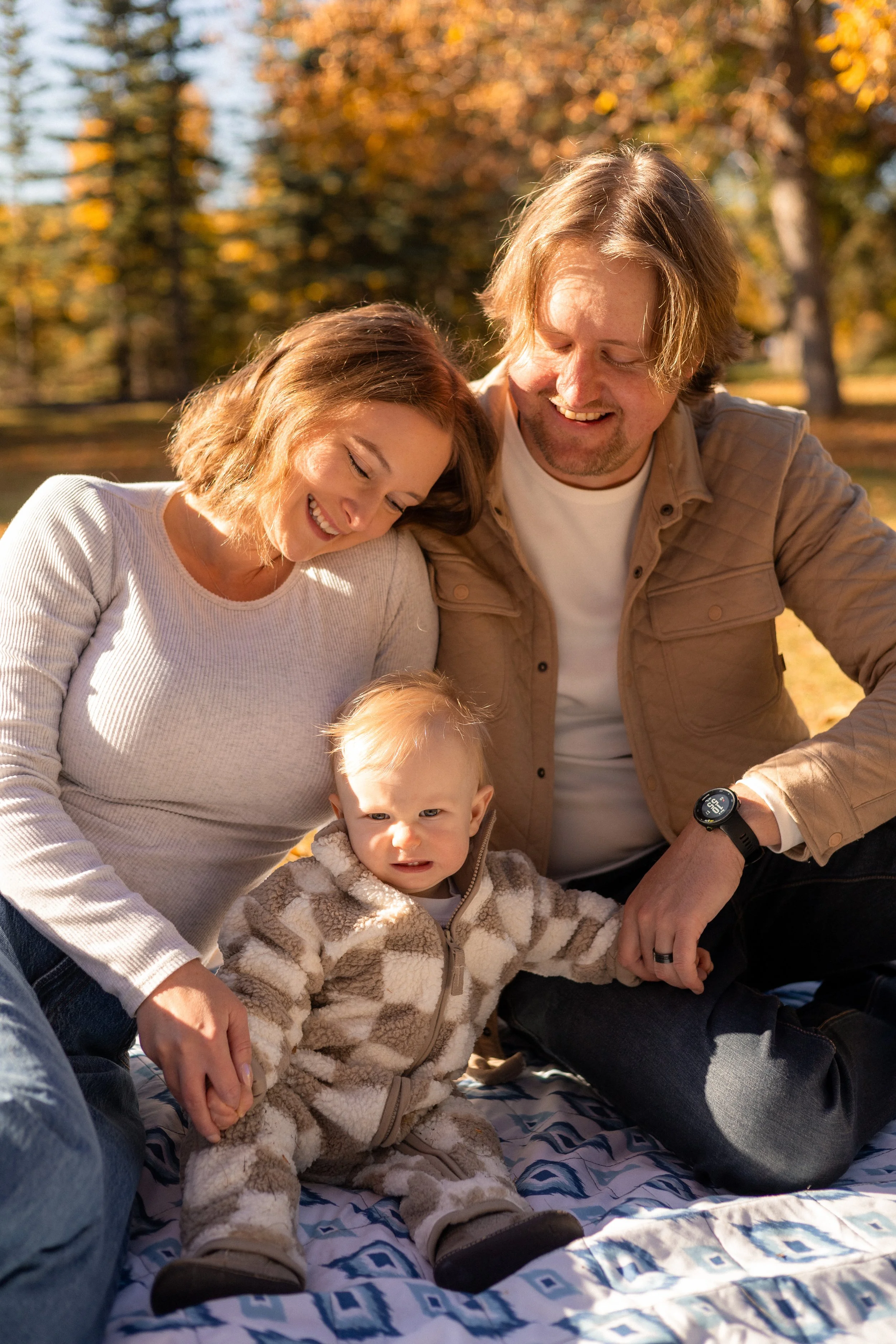 A family of three, including a man and woman with a young child, enjoying a day outdoors during autumn, sitting on a colorful blanket with fall foliage in the background.