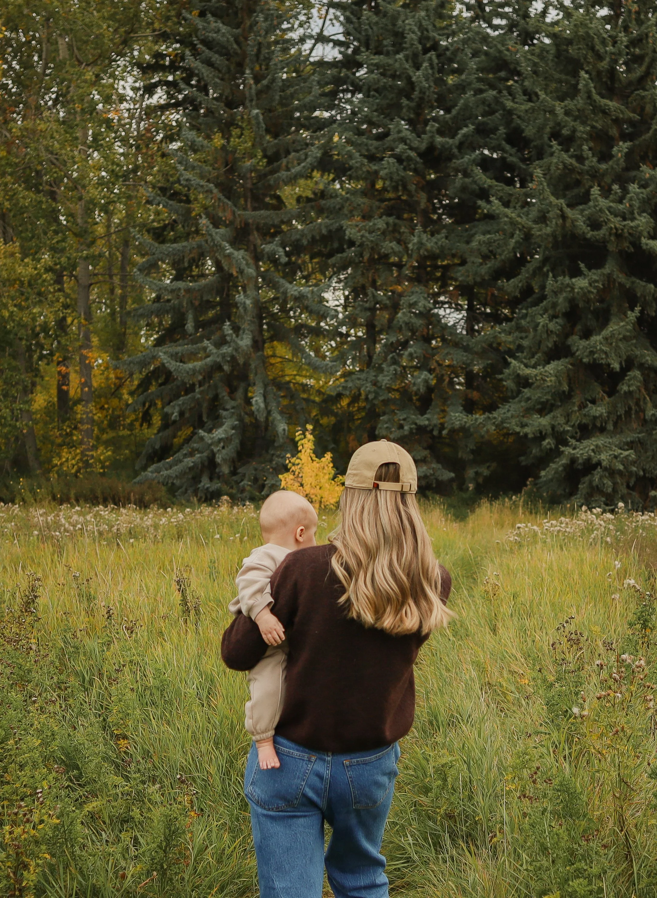 A woman wearing a beige cap and a dark brown sweater holds a young child in a tan hoodie and beige pants. They are standing outdoors in a grassy field surrounded by trees, with large evergreen trees in the background.  Location is Confederation Park 
