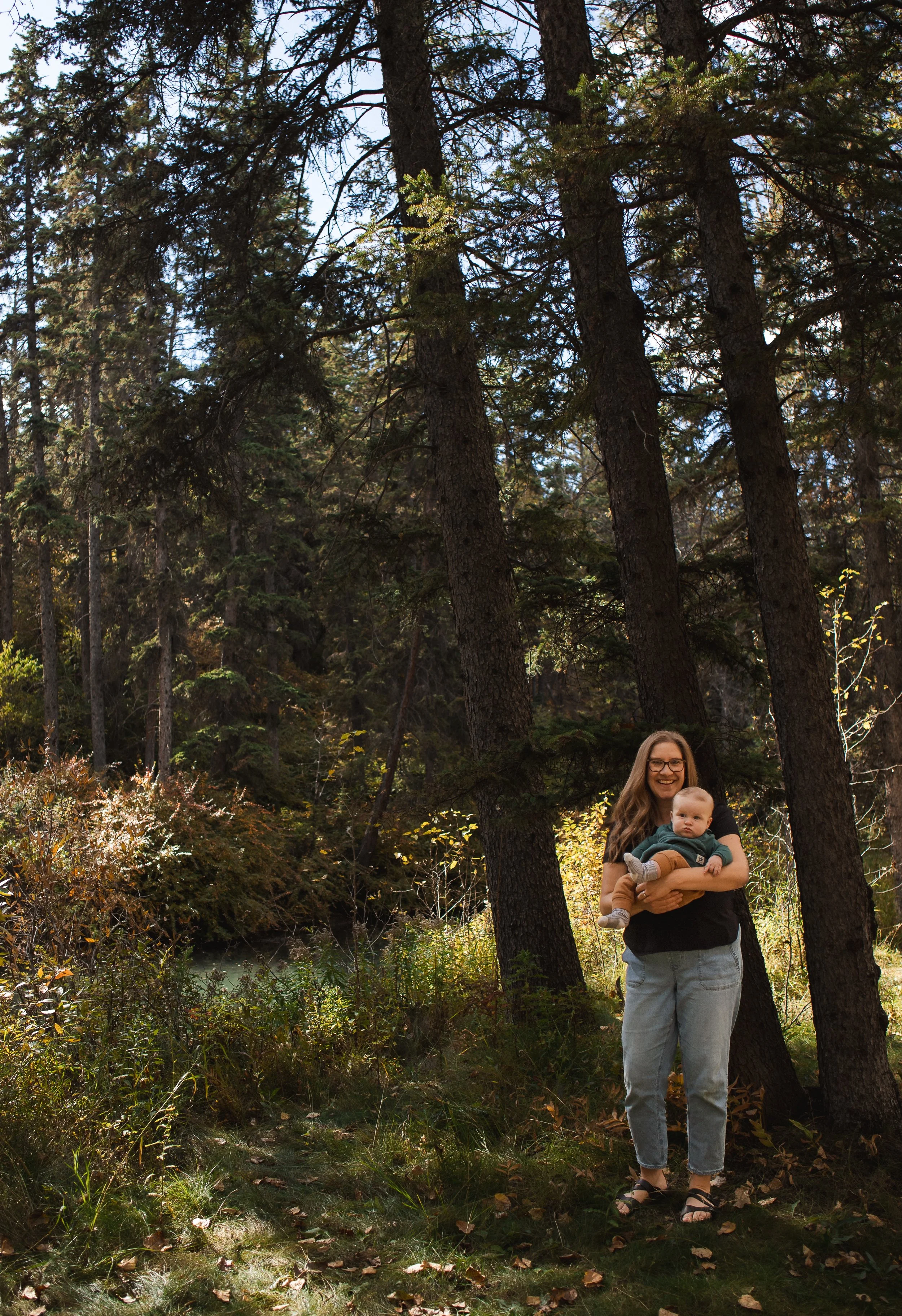 A woman with long brown hair, wearing glasses, a black shirt, and light blue jeans, is holding a baby in a green outfit. They are standing in a forest with tall trees and green foliage in autumn. The woman is smiling and the baby is looking at the ca