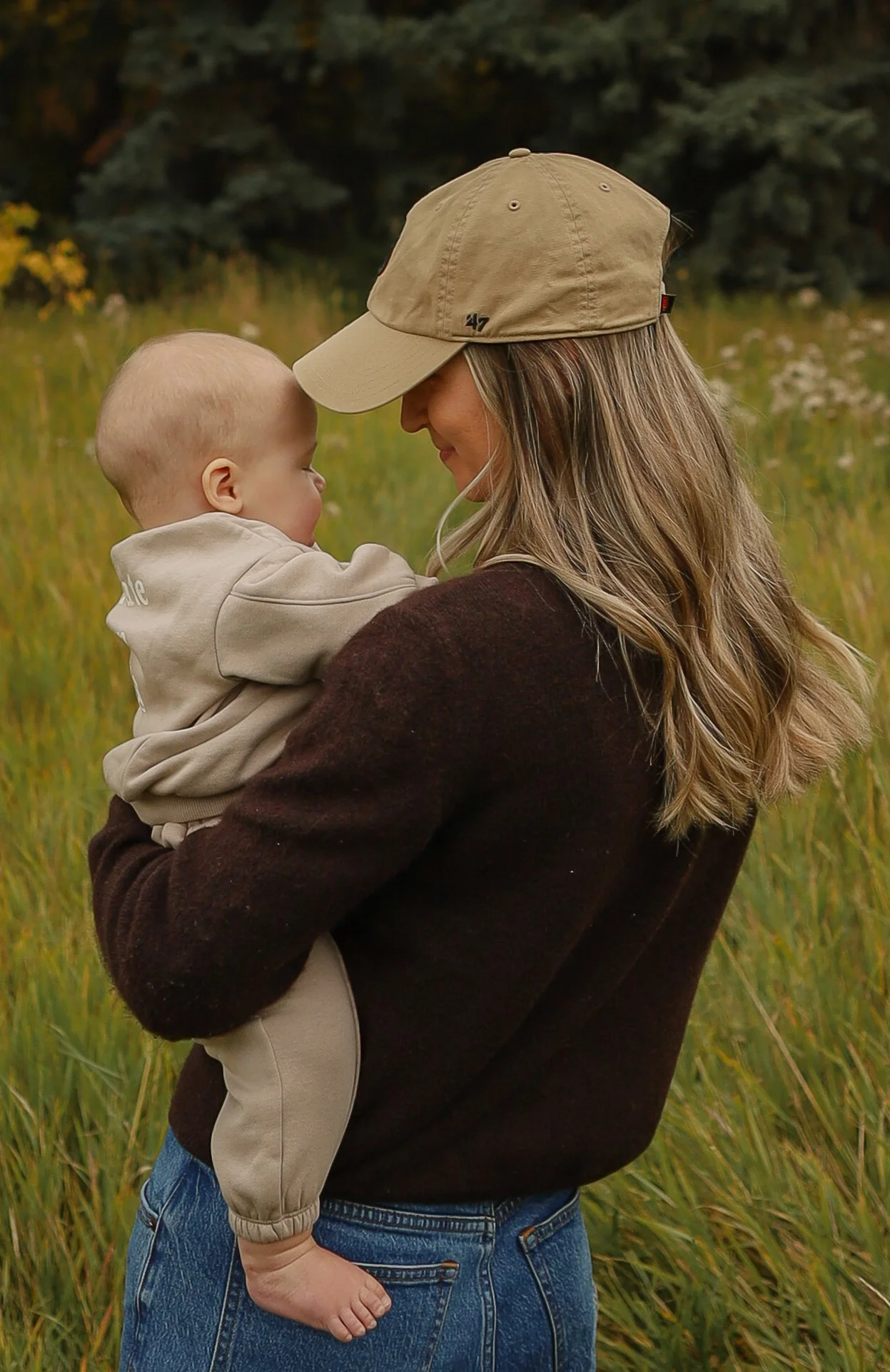 A woman holding a young child in a grassy field during autumn. The woman is wearing a beige cap and a dark sweater, and the child is dressed in a light beige outfit.  Location is Confederation Park in Calgary, Alberta