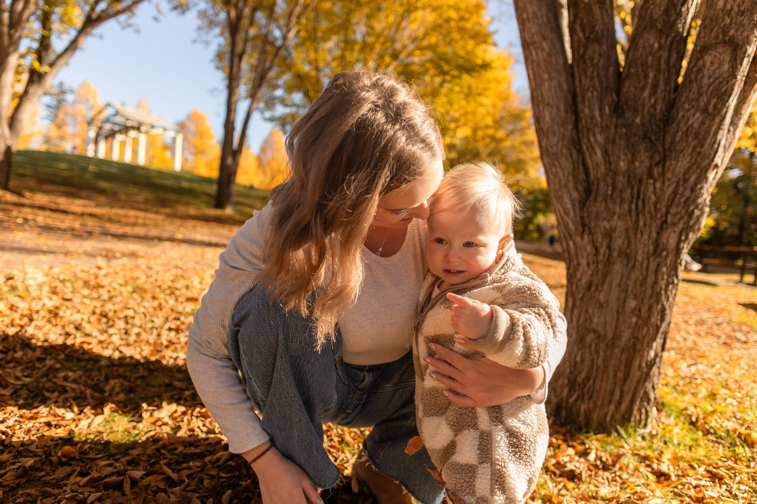 A woman and a young child are outside in a park during autumn, surrounded by colorful fall trees and fallen leaves. The woman is leaning in close to the child, both are smiling, and the child is pointing with one finger.