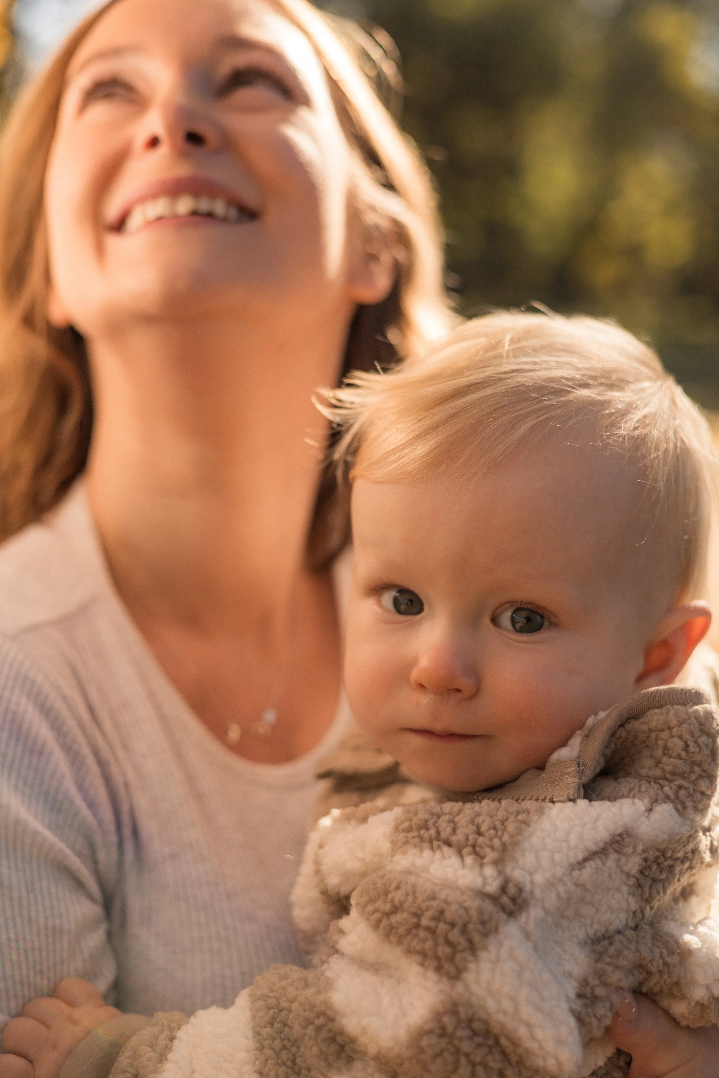 A woman with light brown hair smiling and looking upward, holding a young child with blonde hair and blue eyes, wrapped in a beige and white fluffy blanket, outdoors with a blurred background of trees and sunlight. Location is Baker Park in Calgary, 