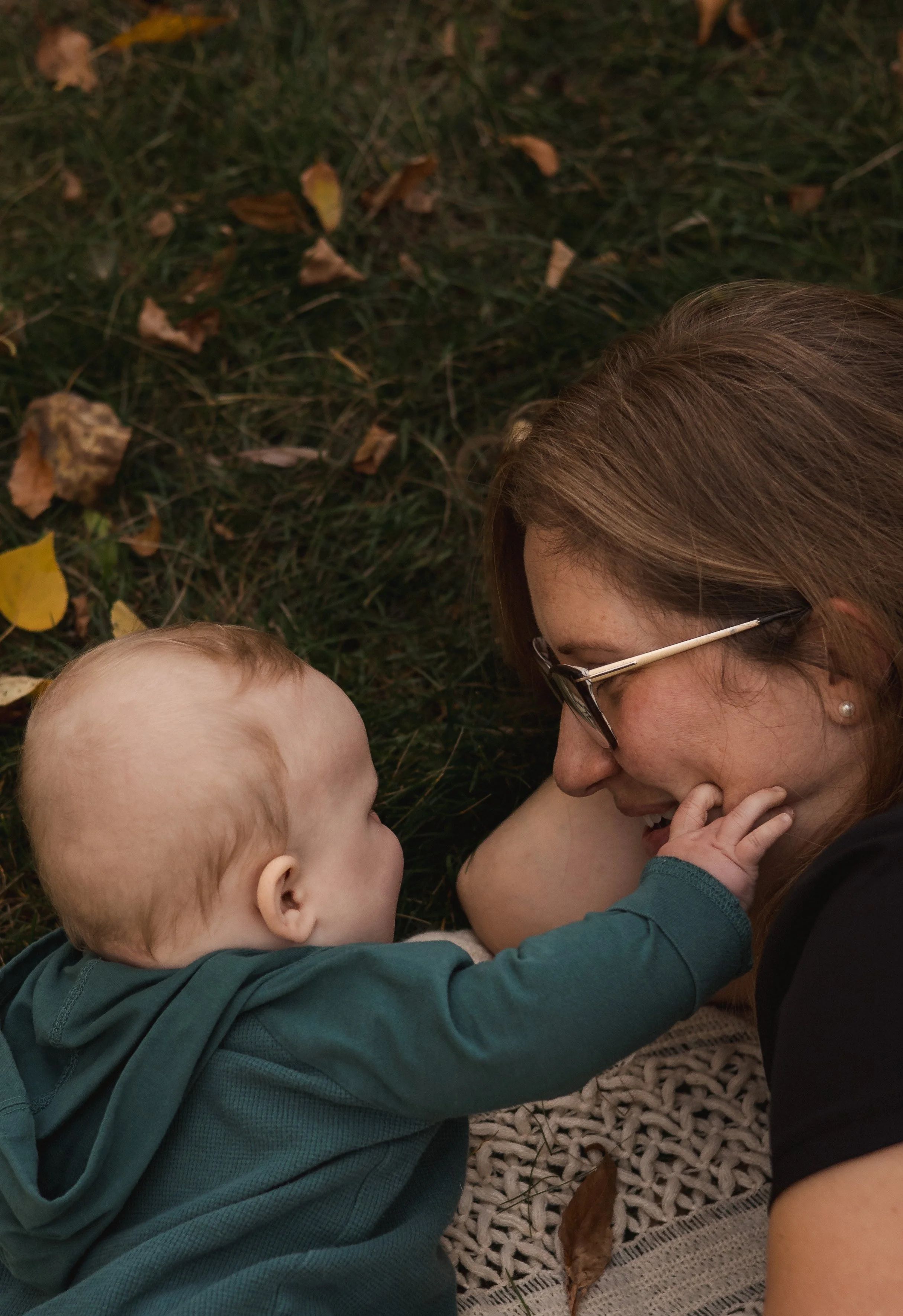 A woman with glasses lying on the ground, smiling and touching the cheek of a baby who is lying on their back, both surrounded by fallen leaves and grass. Location is Bowness Park in Calgary, Alberta