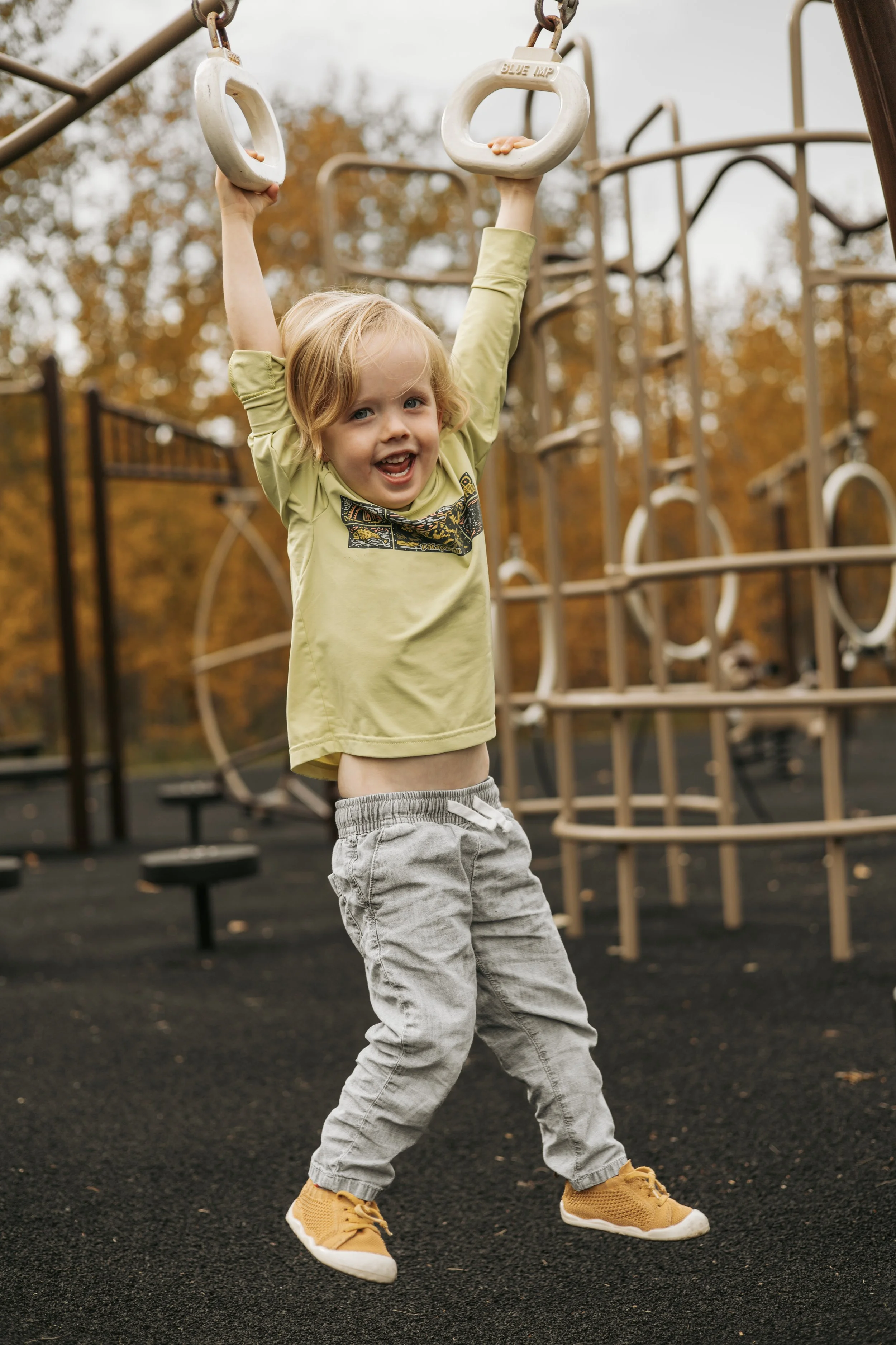 A young child with blond hair wearing a yellow t-shirt, gray pants, and yellow sneakers playing on playground equipment with gymnastic rings, with trees in the background during autumn.