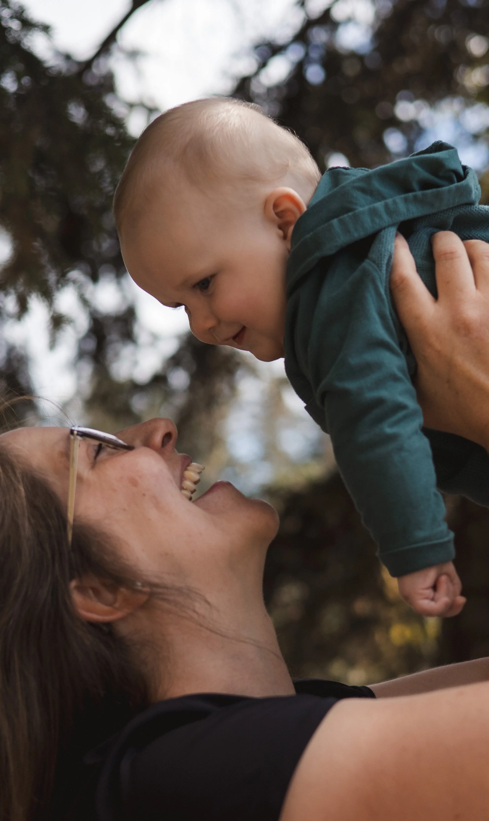 A woman is lying down and holding a young boy up in the air, both smiling as they enjoy a moment outdoors.
