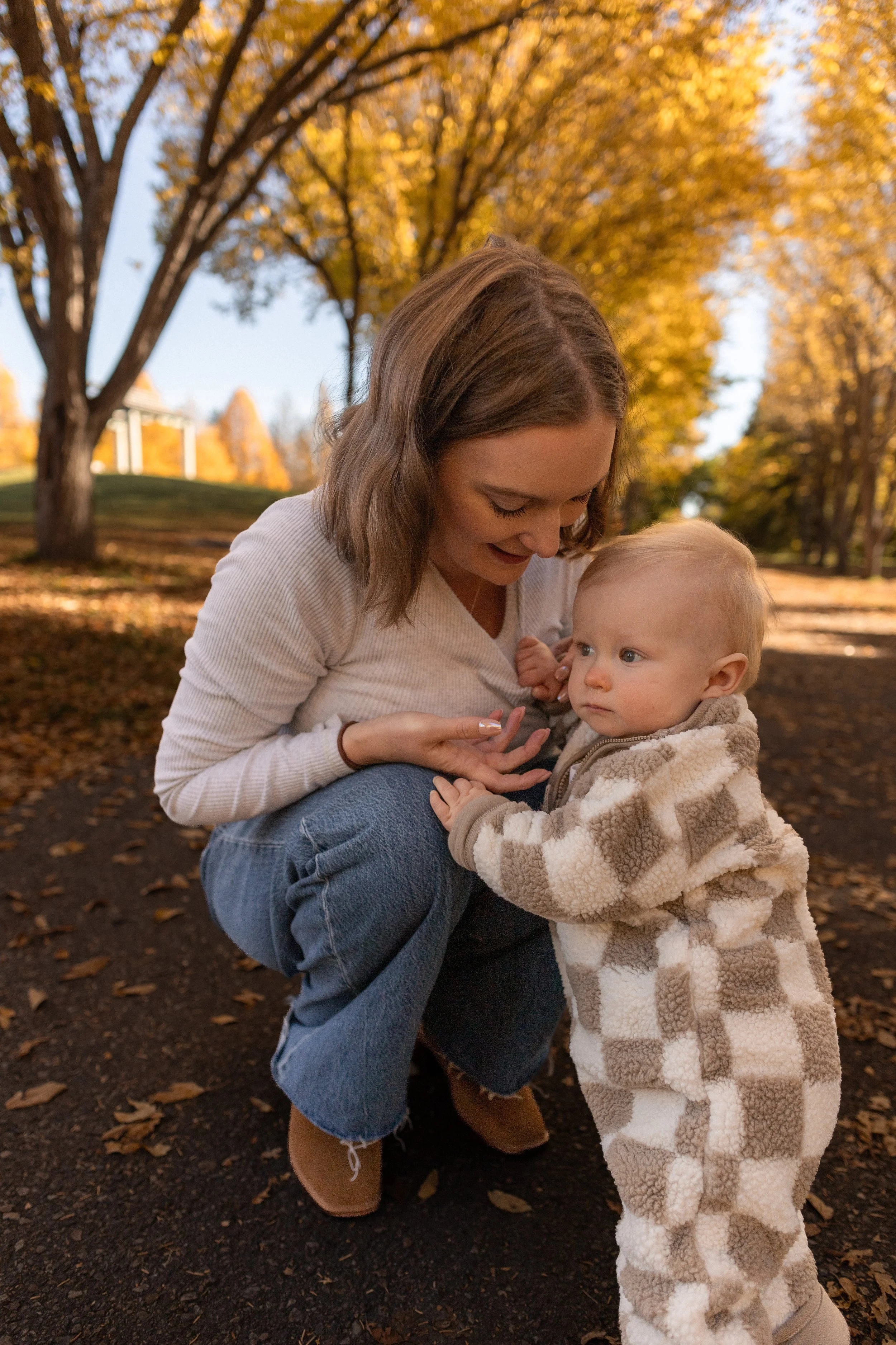 A woman and a young child outdoors in a park during autumn with trees in vibrant fall foliage. The woman is crouching, smiling, and looking at the child. The child is standing in front with a serious expression, wearing a beige and white striped flee