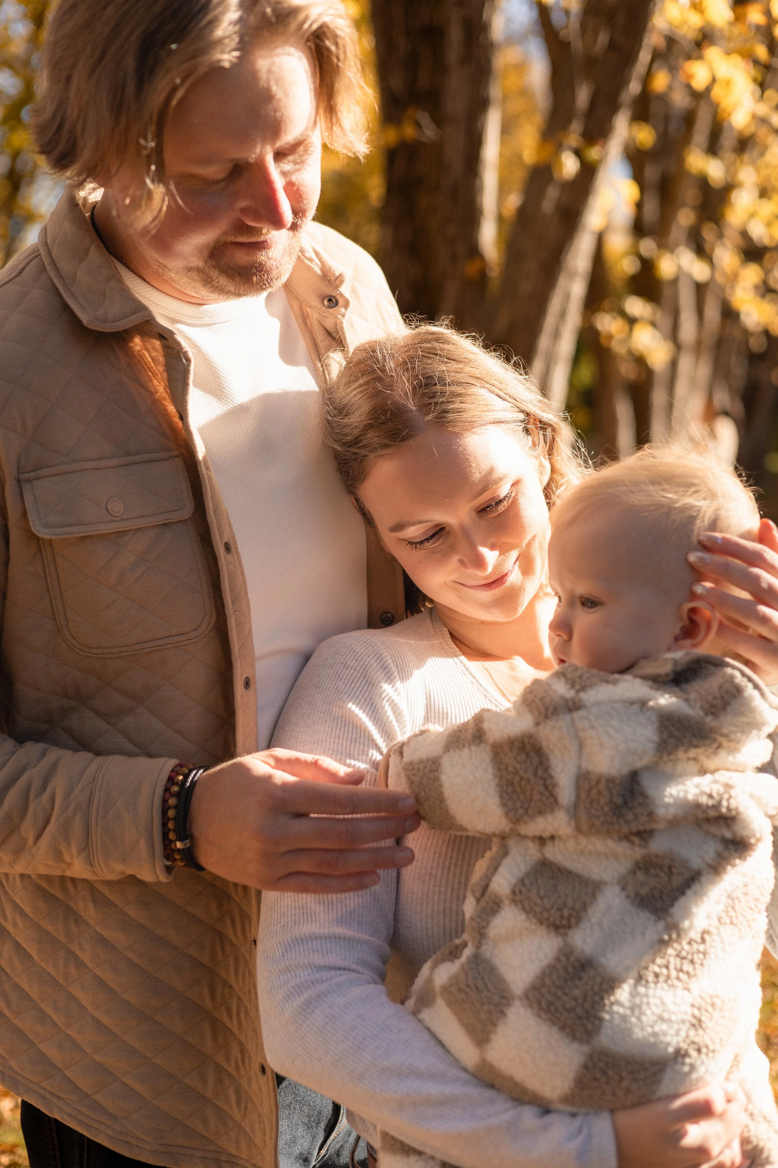 A family scene outdoors with a man, woman, and young child. The man has shoulder-length brown hair, a beard, and is wearing a beige jacket. The woman has light brown hair tied back, and is wearing a white shirt. The child has blonde hair and is dress