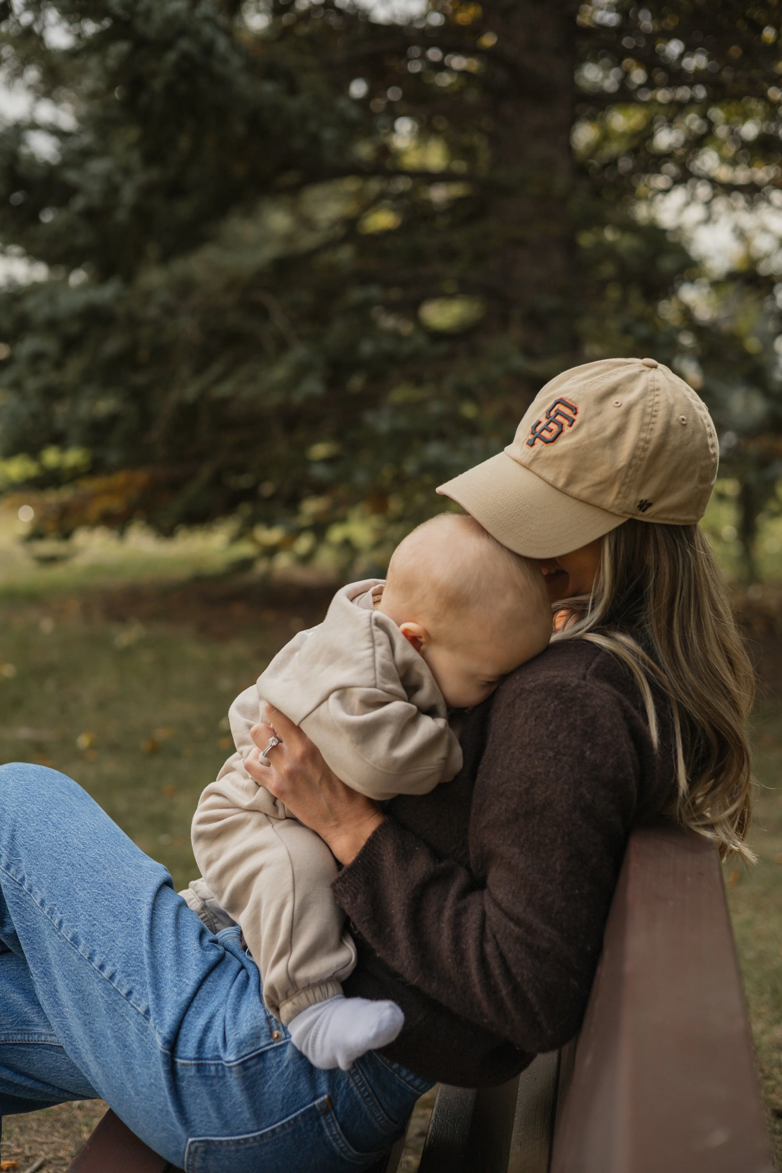 A woman wearing a beige cap with a logo on it, sitting on a park bench, holds a young child in her arms, both sharing a hug. They are outdoors with trees and greenery in the background.  Location is Confederation Park in Calgary, Alberta