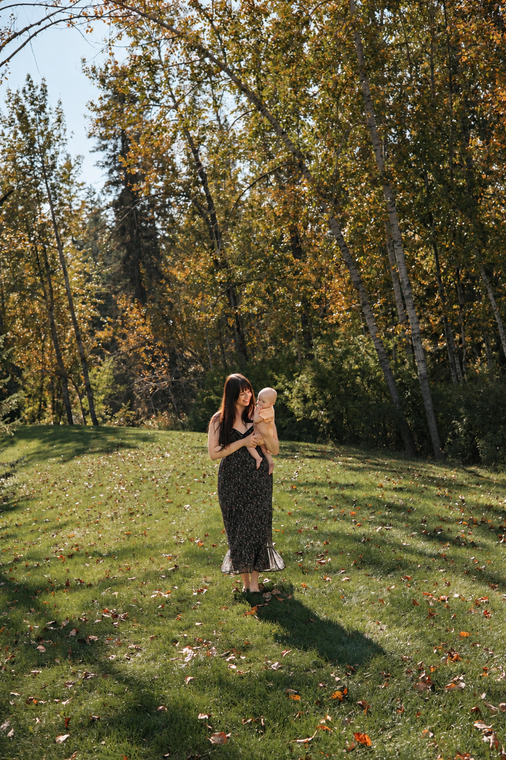 A woman in a black floral dress holding a baby outside on a sunny autumn day with trees and fallen leaves in the background.