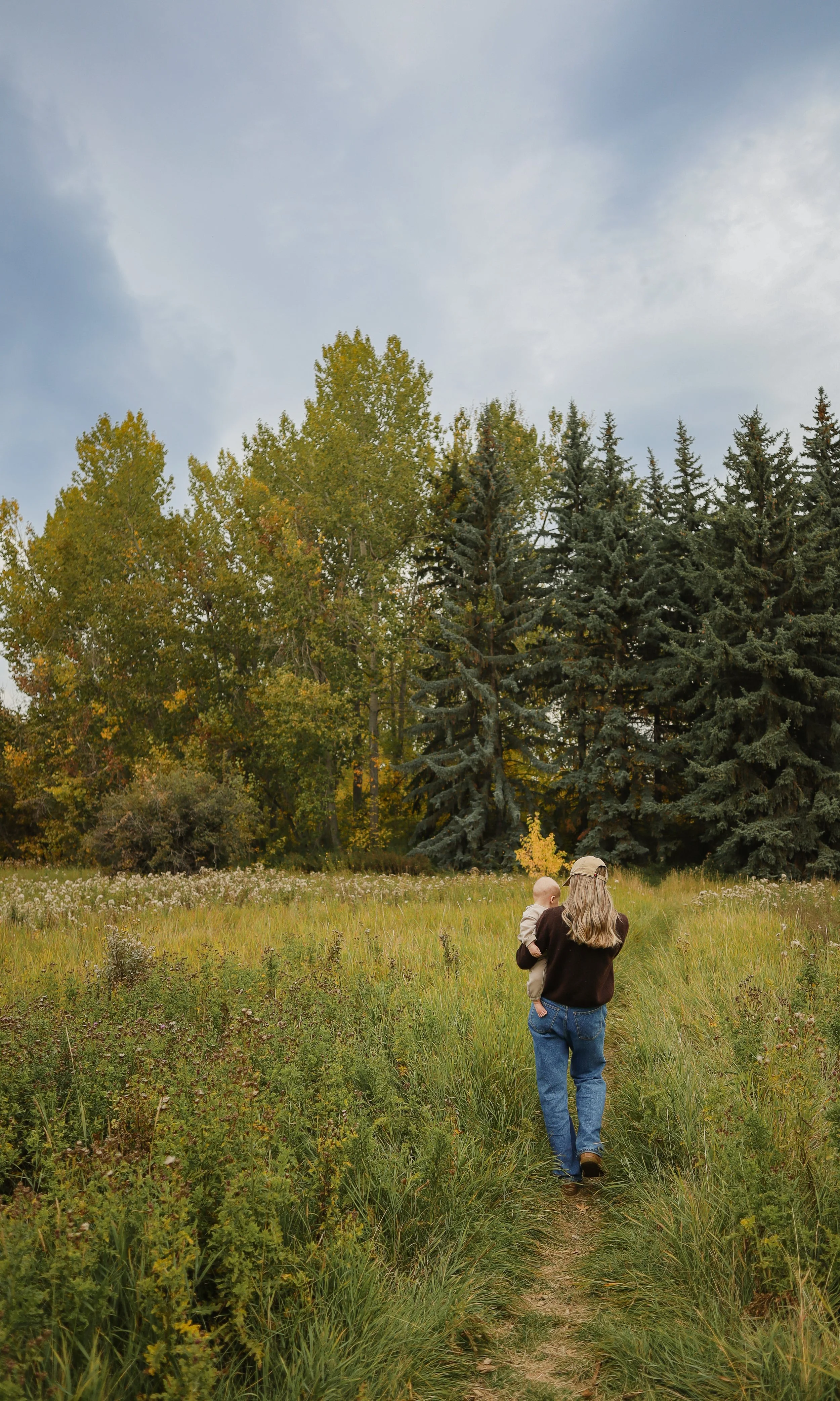A woman walking on a grassy trail while holding a child, surrounded by tall green trees and an overcast sky.  Location is Confederation Park in Calgary, Alberta