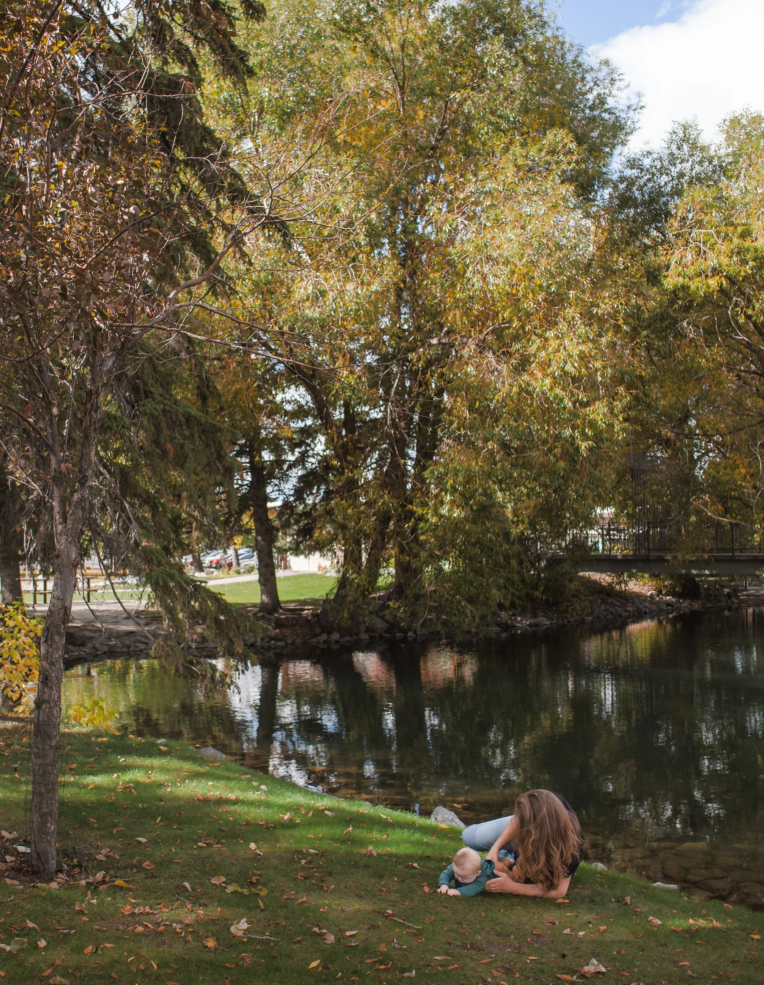 A woman with long brown hair lying on the grass next to a small child by a pond in a park, with trees and a bridge in the background.