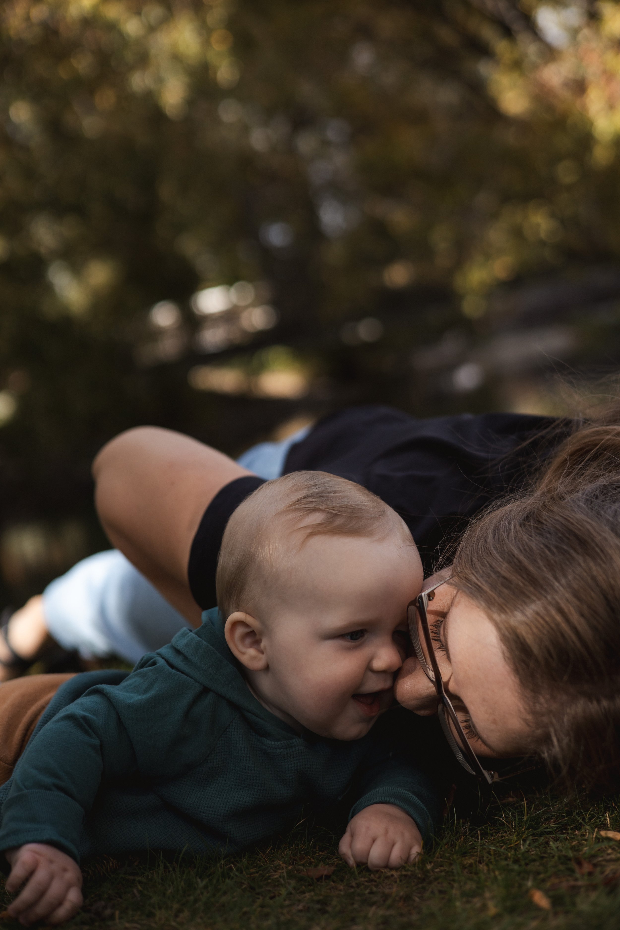 A young child with blond hair and a woman with glasses and brown hair are laying on the grass outdoors, close together, with the child's face near the woman's nose. The background is blurry with trees and sunlight. Location is Bowness Park in Calgary