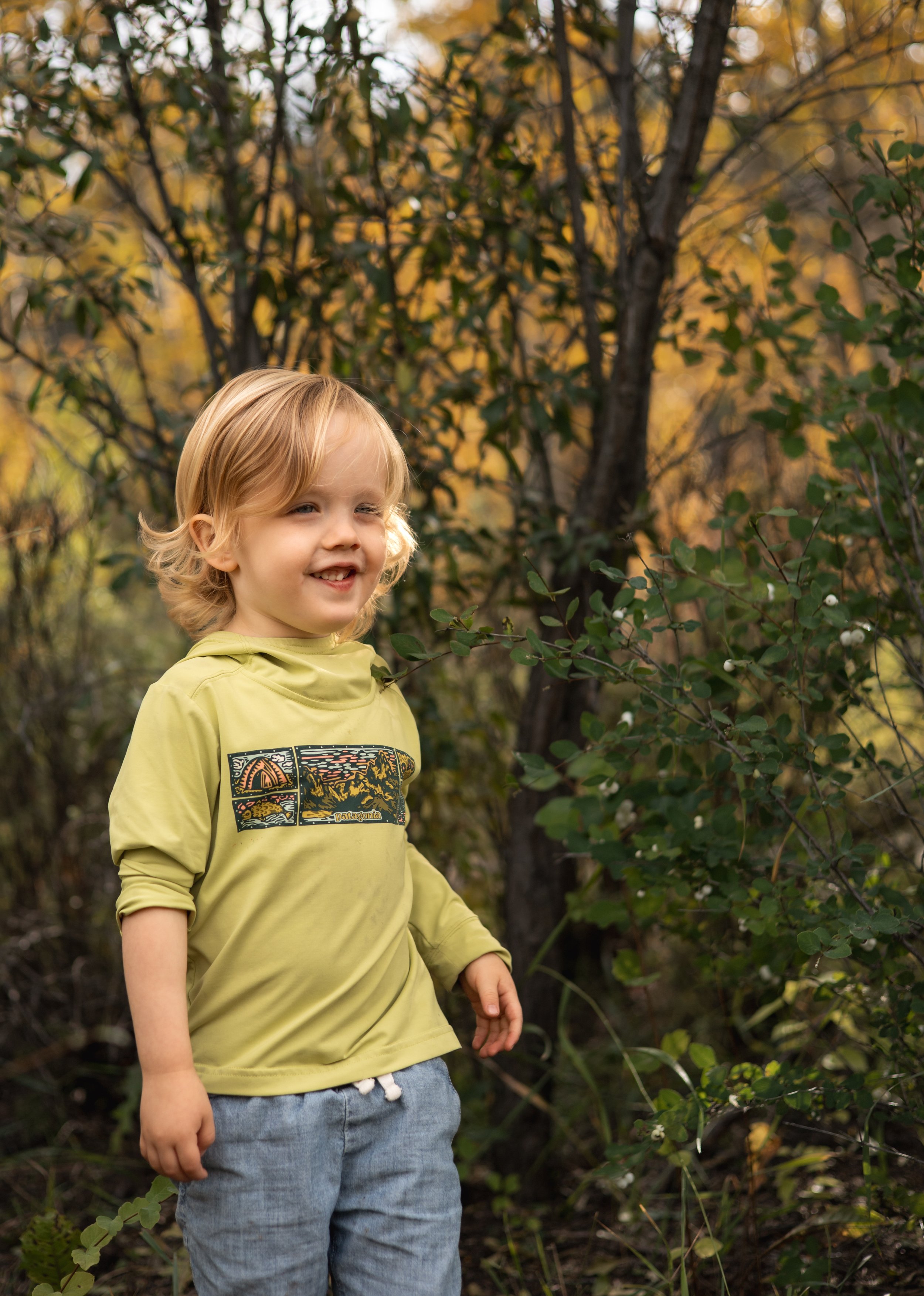 A smiling young child with blonde curly hair wearing a yellow hoodie with a graphic and blue jeans, standing outdoors among green bushes with a background of autumn-colored trees.
