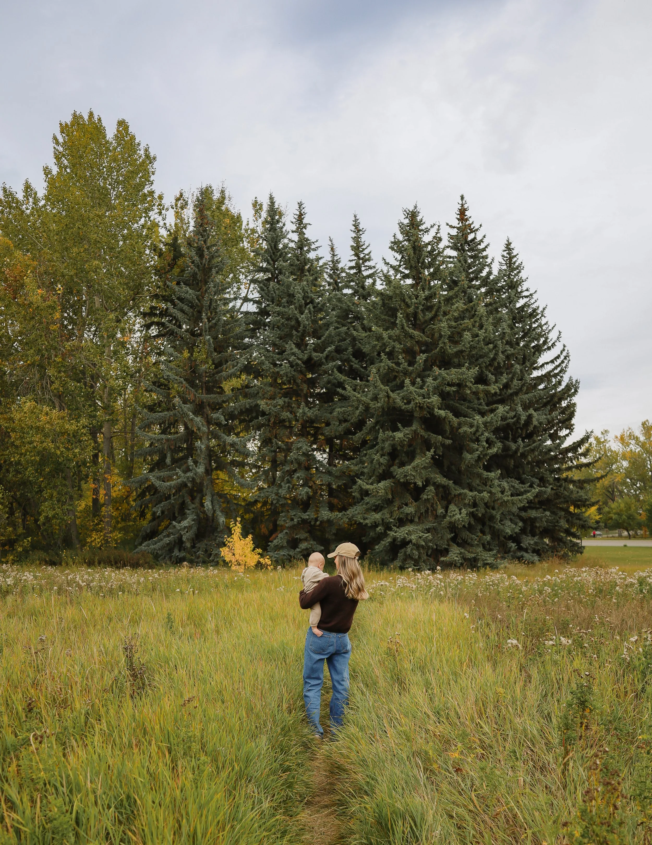 A woman holding a child in front of large pine trees in a grassy park or field.