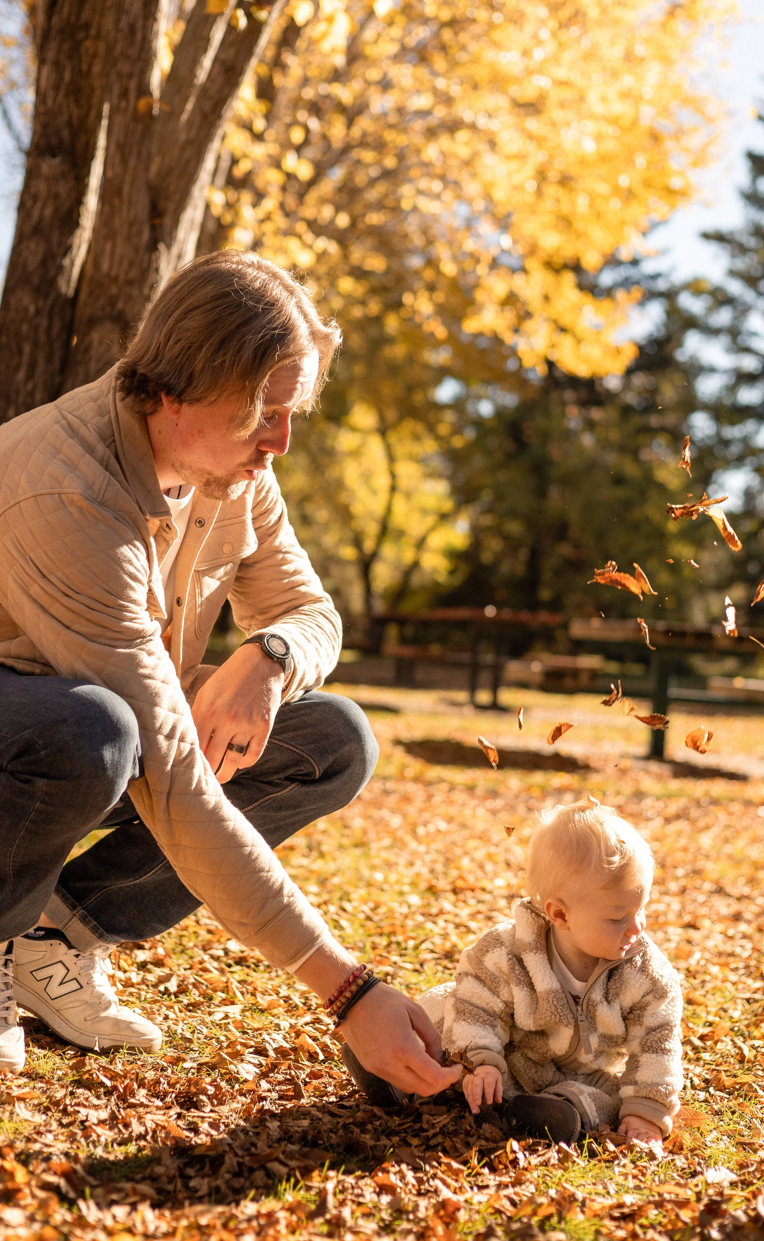 A man crouches in a park during fall, playing with a young child on a blanket covered with autumn leaves, with trees showing yellow and orange foliage in the background.
