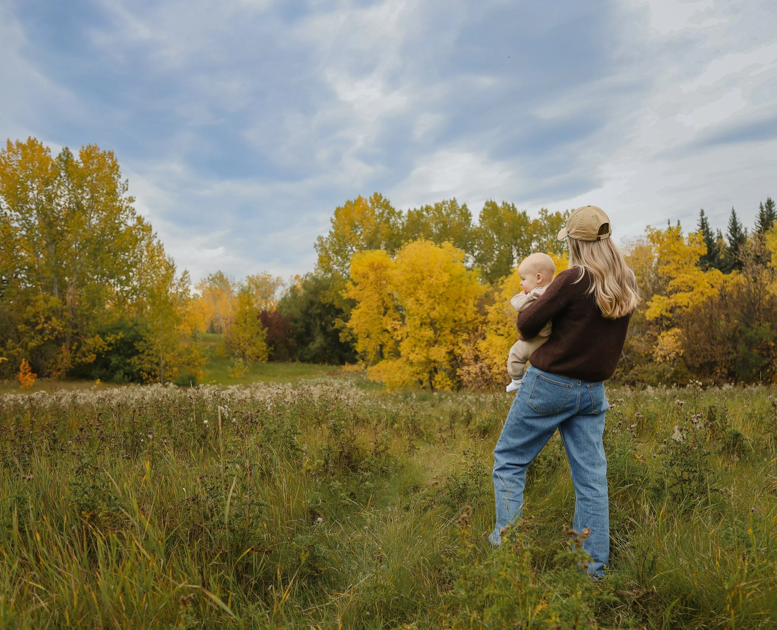 A woman holding a baby in a park with trees showing autumn colors under a partly cloudy sky.  Location is Confederation Park in Calgary, Alberta