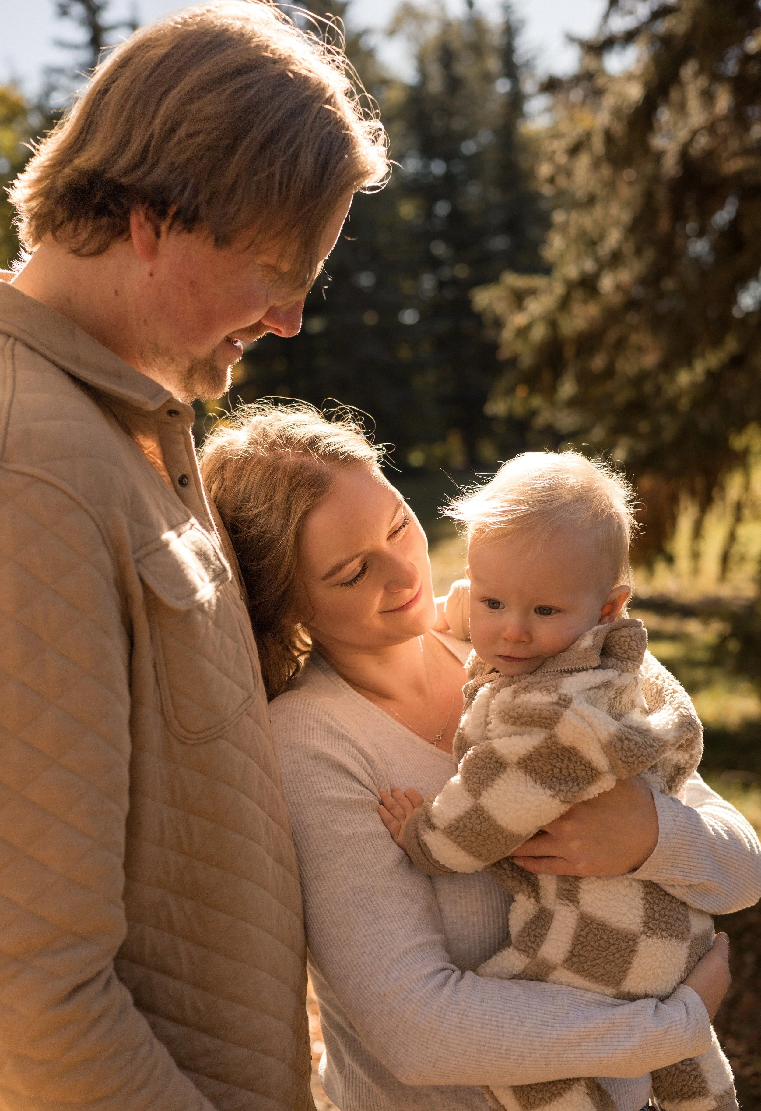 A family of three outdoors during golden hour. The father, with light brown hair and beige quilted jacket, is smiling while looking down at the toddler. The mother, with long brown hair and a white top, is holding the young child, who has light blond