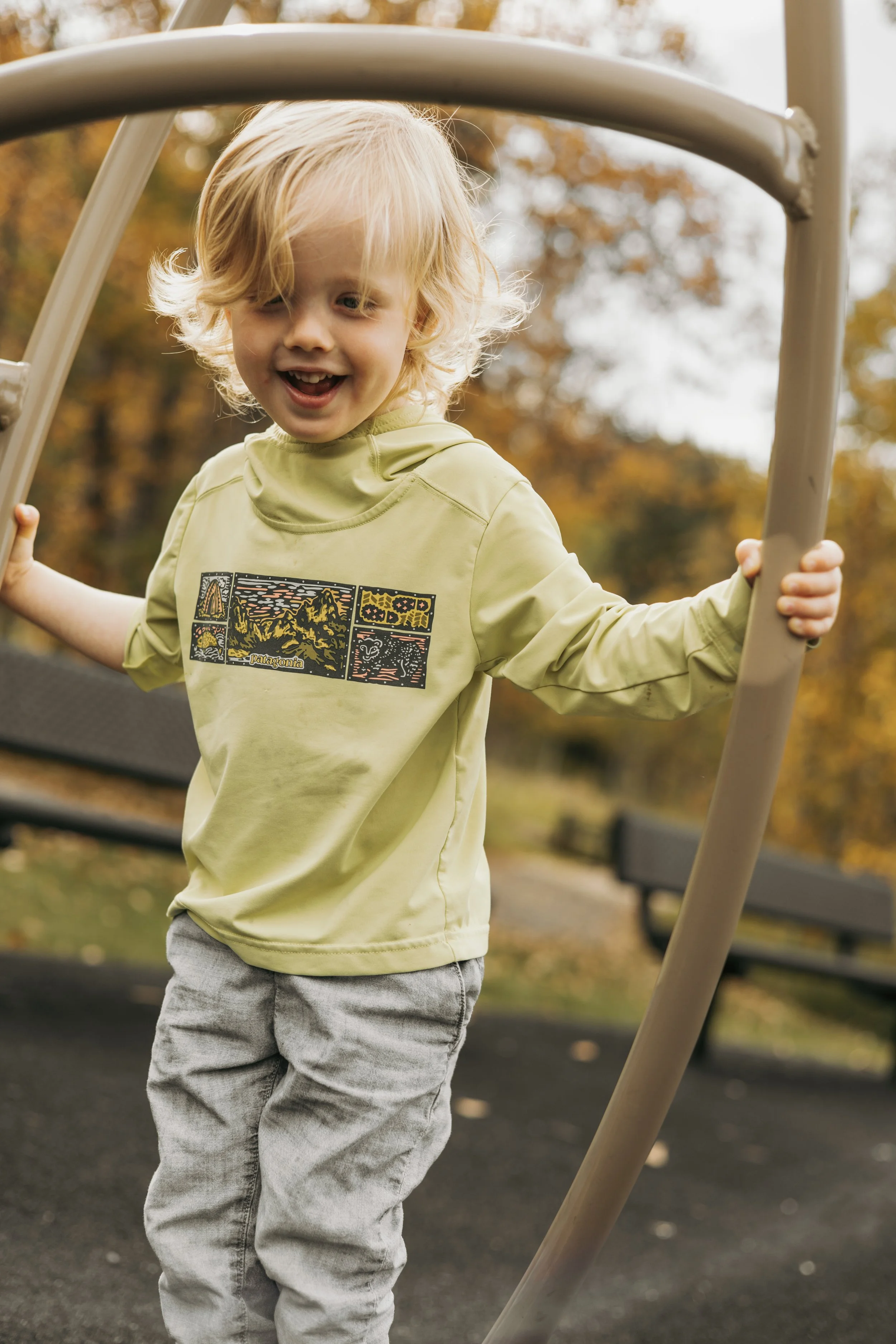 A young blonde-haired boy smiling and playing on a playground slide outdoors during fall, wearing a yellow long-sleeve shirt and gray pants, with trees in the background.
