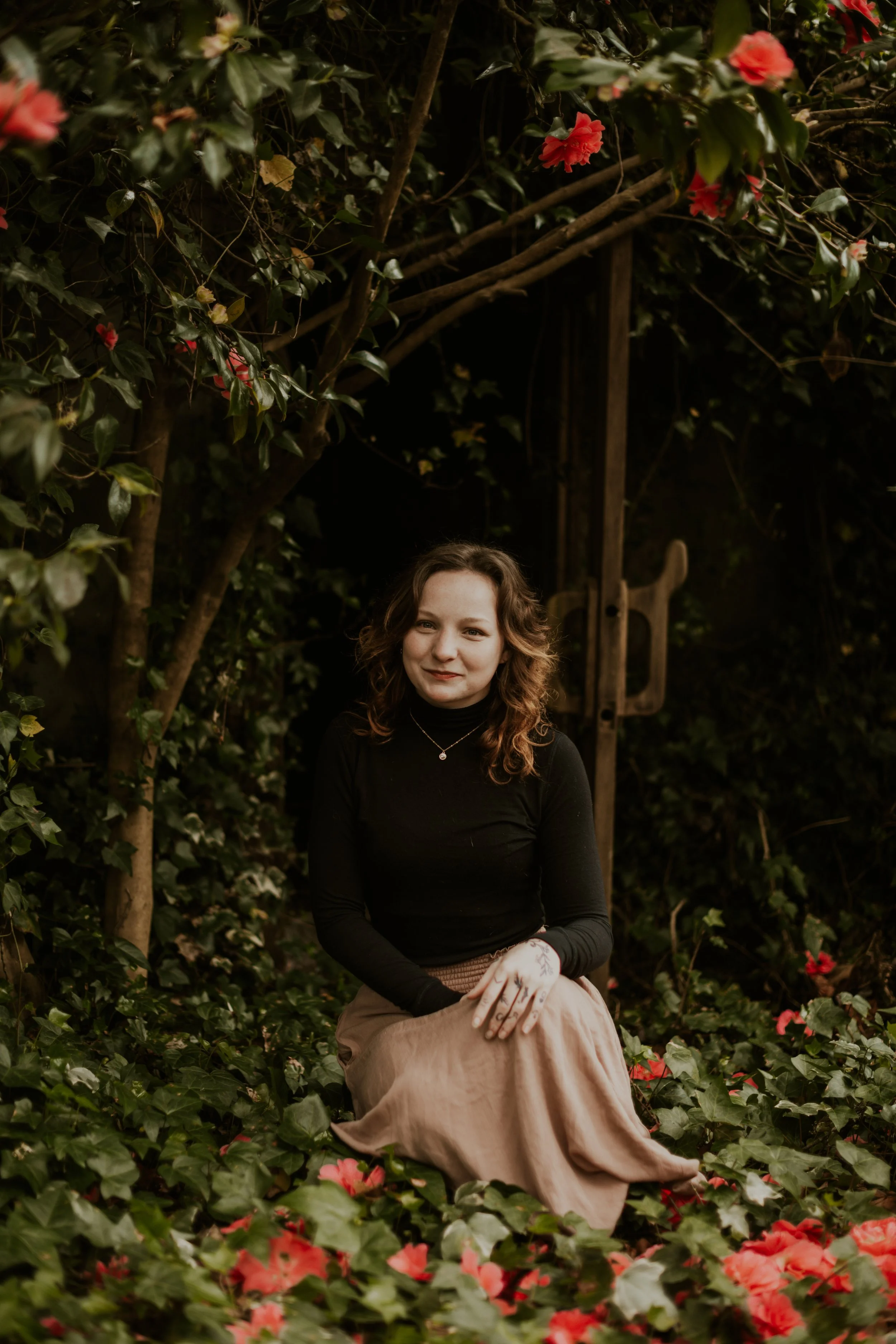 Vera Lee Bird in a black shirt and pink skirt in front of a garden door surrounded by leaves and flowers with a small golden necklace and warm smile