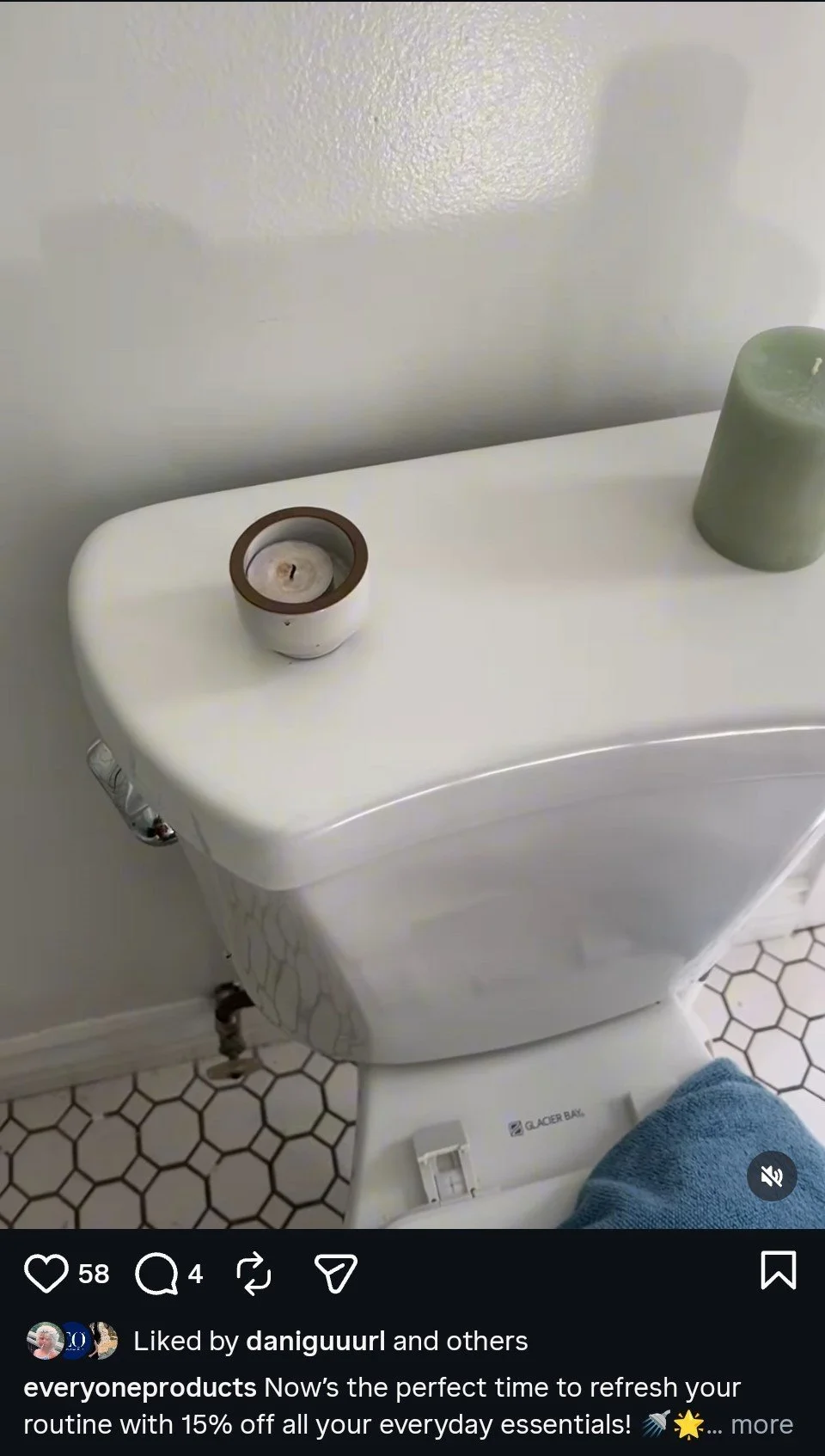 A white toilet with a blue towel hanging on the side, a green candle on the tank, and a small candle holder on top of the toilet tank. The bathroom has white walls and hexagonal black-and-white floor tiles.