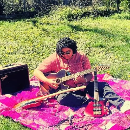 A person sitting on a pink blanket outdoors, playing an acoustic guitar, surrounded by musical equipment including an amplifier, a bass guitar, and a microphone, with trees and grass in the background.