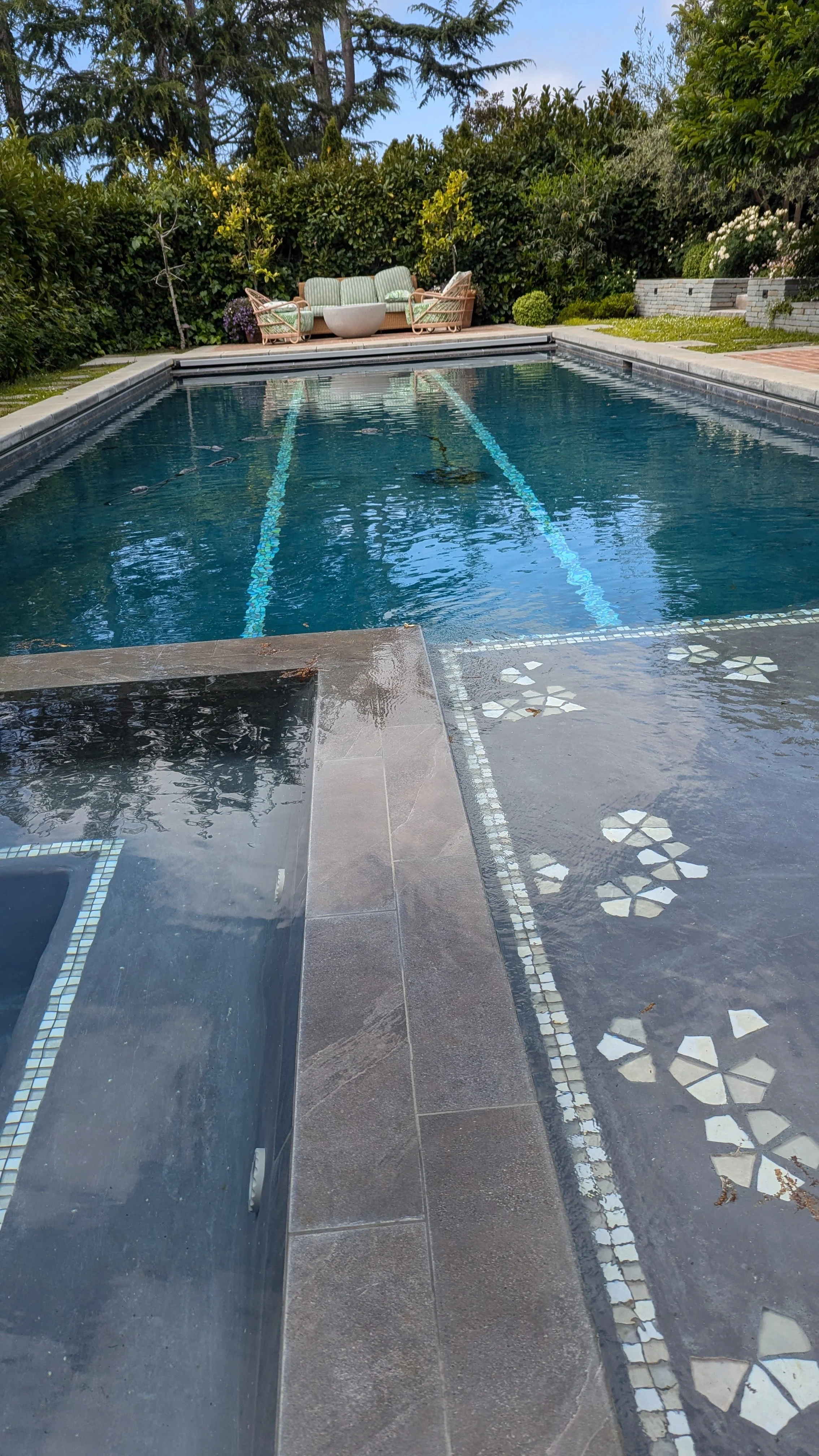 Backyard swimming pool with mosaic tile accents, cushioned patio seating, lush hedges, and landscaped garden