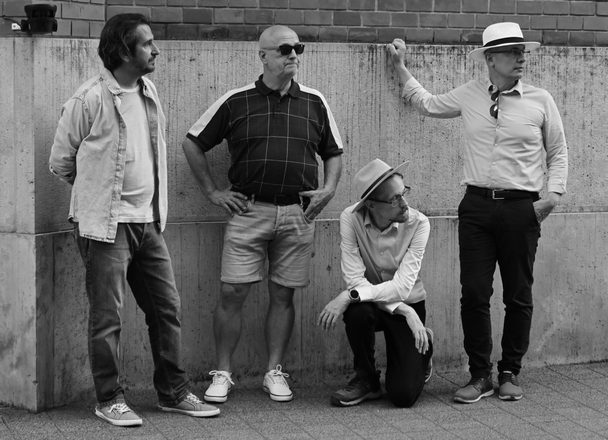 Four men standing against a concrete wall with a brick building behind them, in black and white. One man is seated down, wearing eyeglasses and a hat. The others are standing, two wearing casual clothes, and one wearing sunglasses and a checked polo shirt. One of the standing men is touching the wall with his arm.