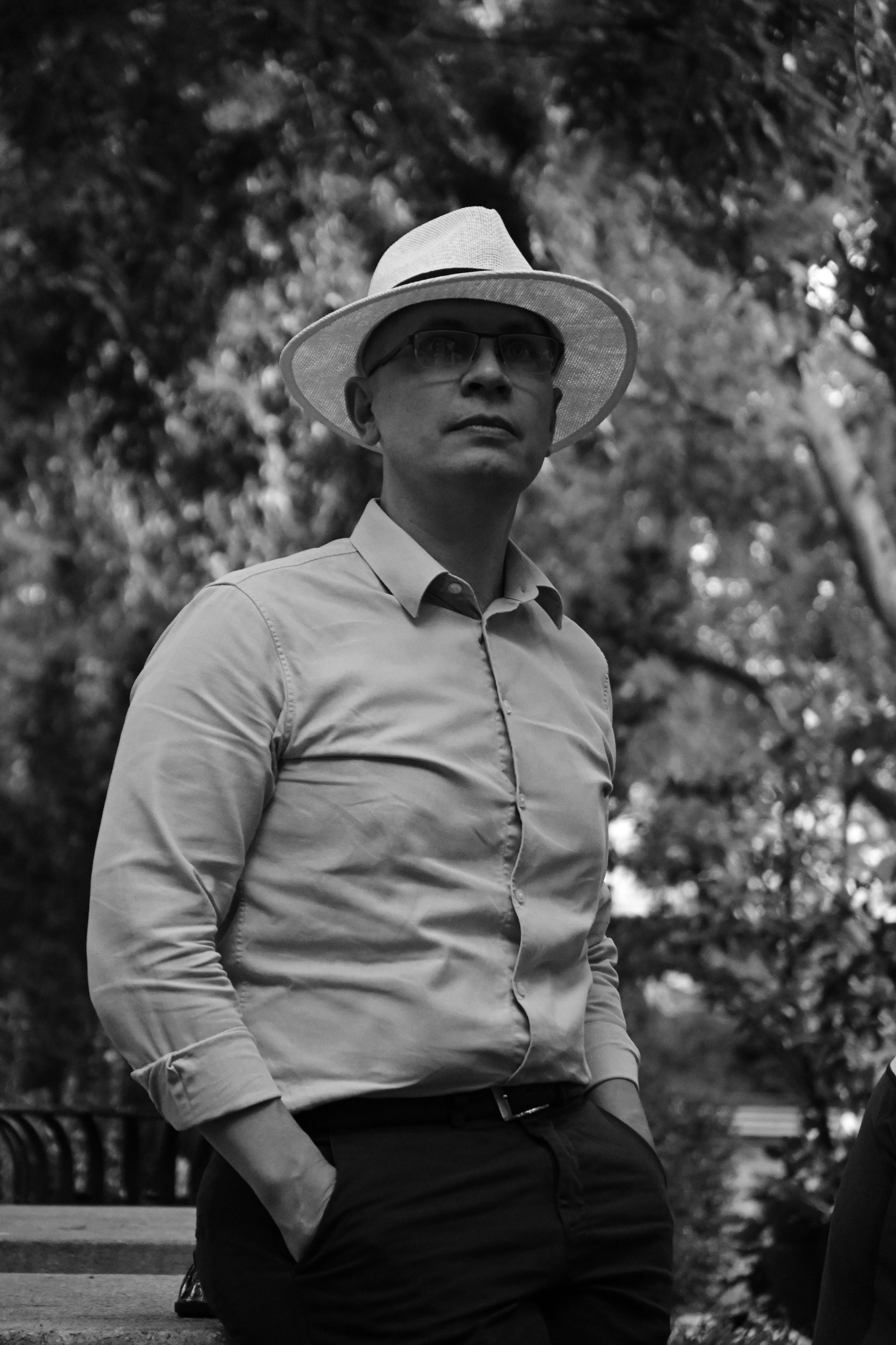 A black and white photo of a man wearing glasses, a hat, a collared shirt, and dark pants, standing outdoors with trees in the background.