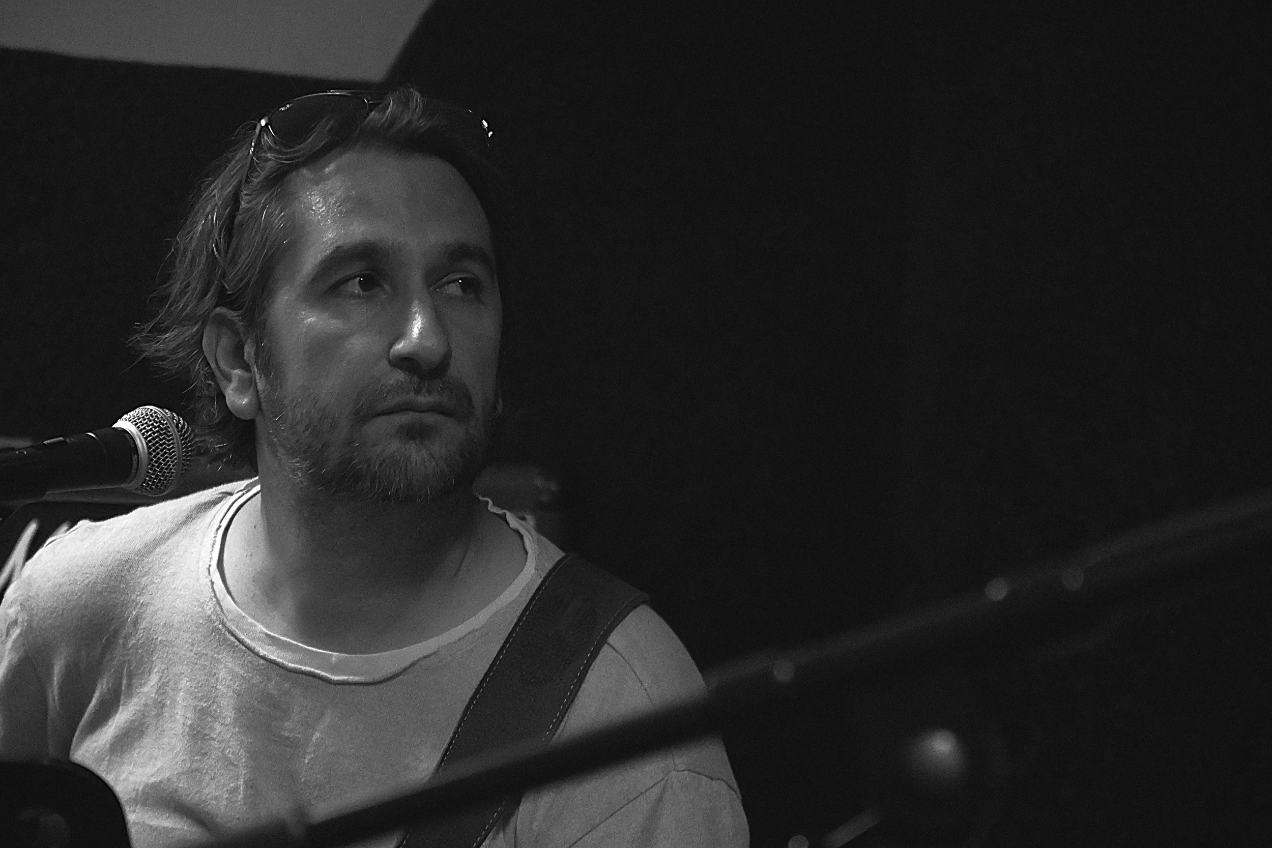 A black and white close-up photo of a man with medium-length hair and a beard, wearing a T-shirt, sitting in front of a microphone with glasses resting on his head.
