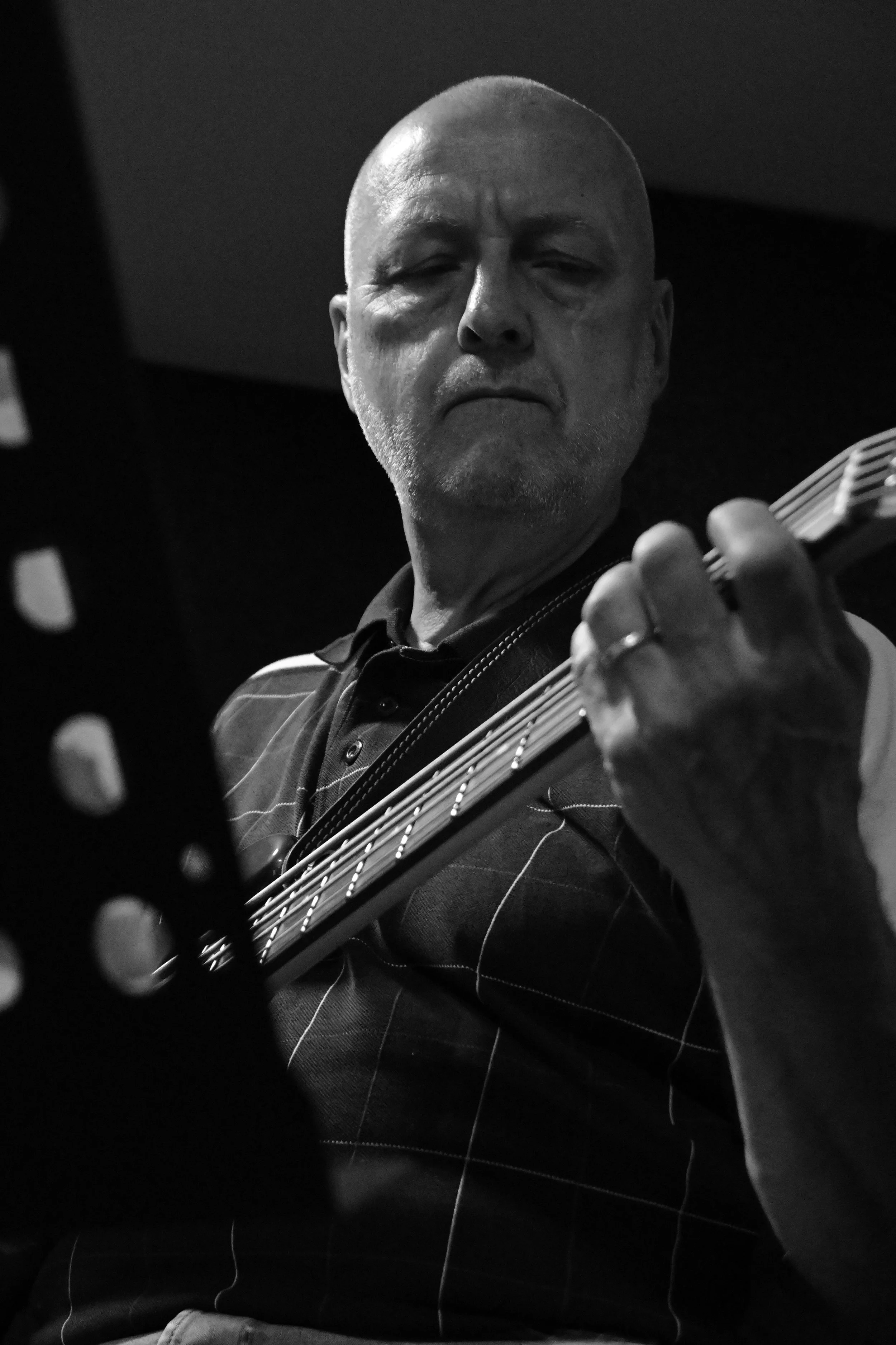 A black and white photo of a man with a bald head and a goatee, holding and playing an electric guitar. The man has a serious expression and is wearing a dark shirt with white pinstripes.