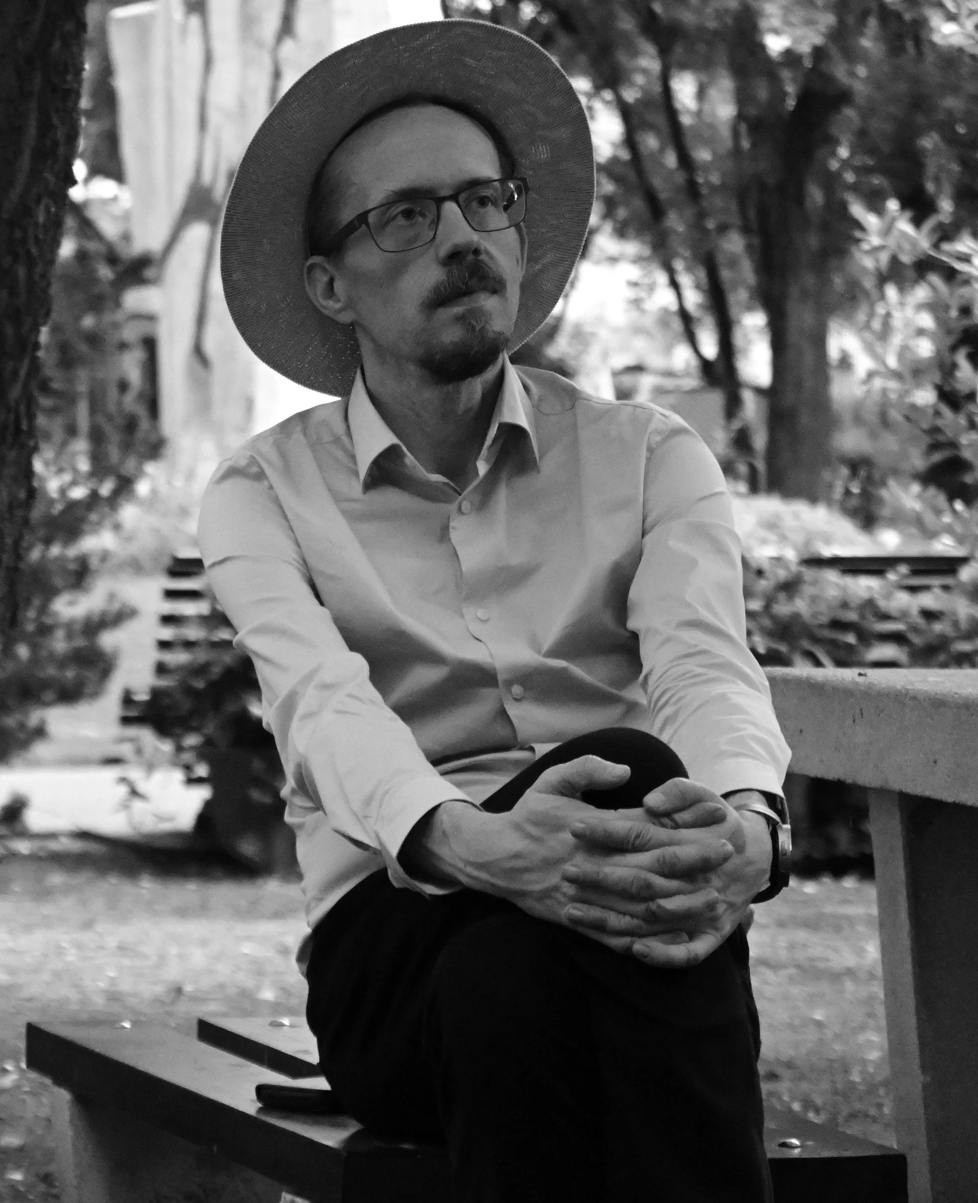 A man wearing glasses, a wide-brimmed hat, and a button-up shirt, sitting outdoors on a park bench with trees in the background. The photo is in black and white.