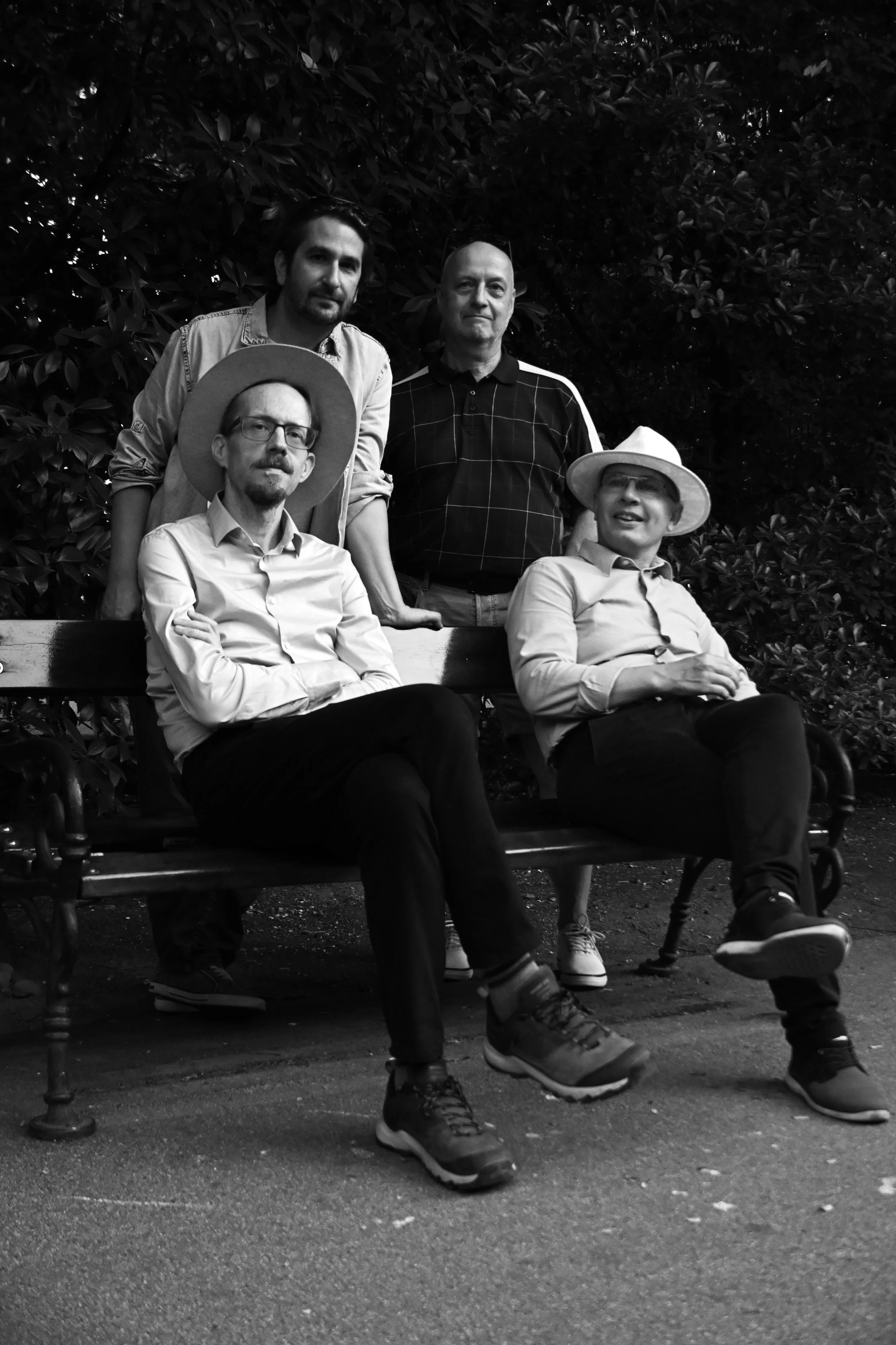 Black and white photo of four men seated on a bench with one standing behind them, in front of bushes. The men are casually dressed, some wearing hats. The man in front left wears glasses and a hat, while the man on the right is wearing glasses and a hat, smiling and looking slightly to the right. The two men standing behind are looking at the camera, one with a serious expression and the other with a neutral expression.