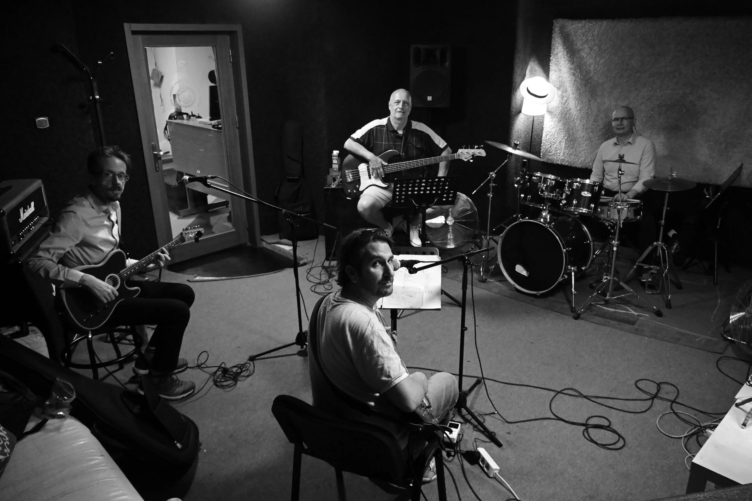 A group of four men in a recording studio, with a drum set, guitar, microphone, and music stand.