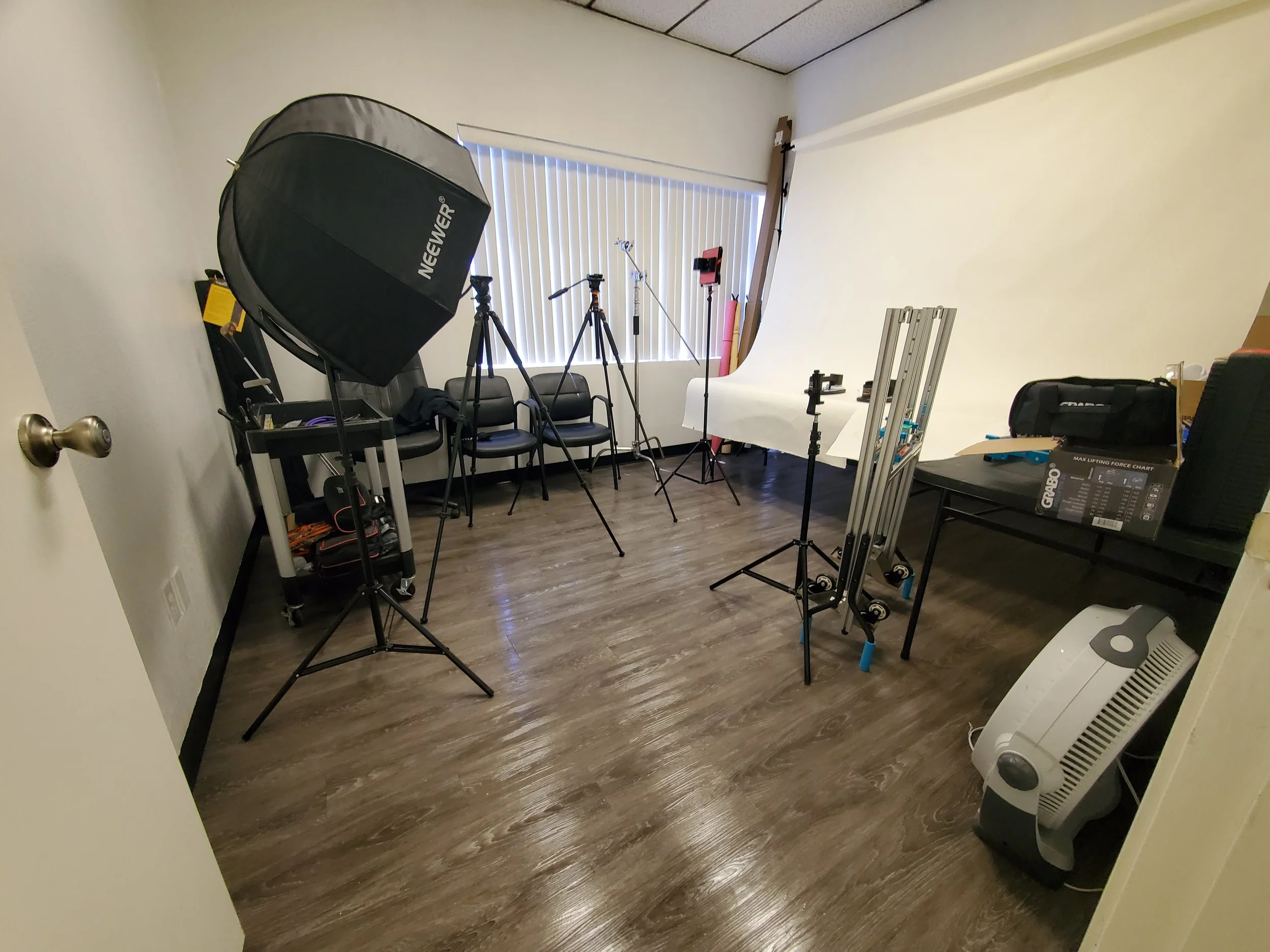 Photography studio with lighting equipment, tripods, chairs, and a white backdrop, in a room with wood flooring and a window with blinds.