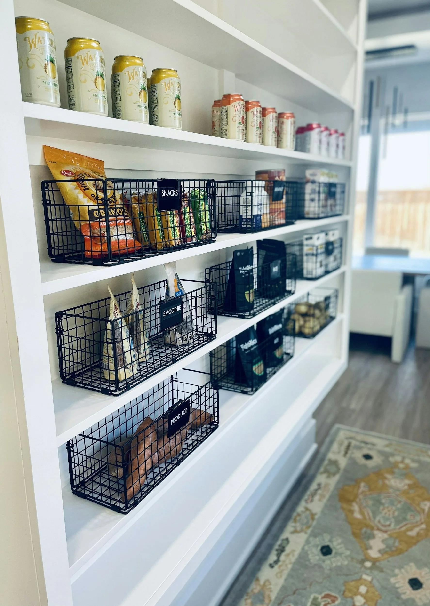 White shelving unit with black wire baskets labeled for snacks, smoothies, produce, and other categories, containing various food and drink items, with a patterned rug on the floor.