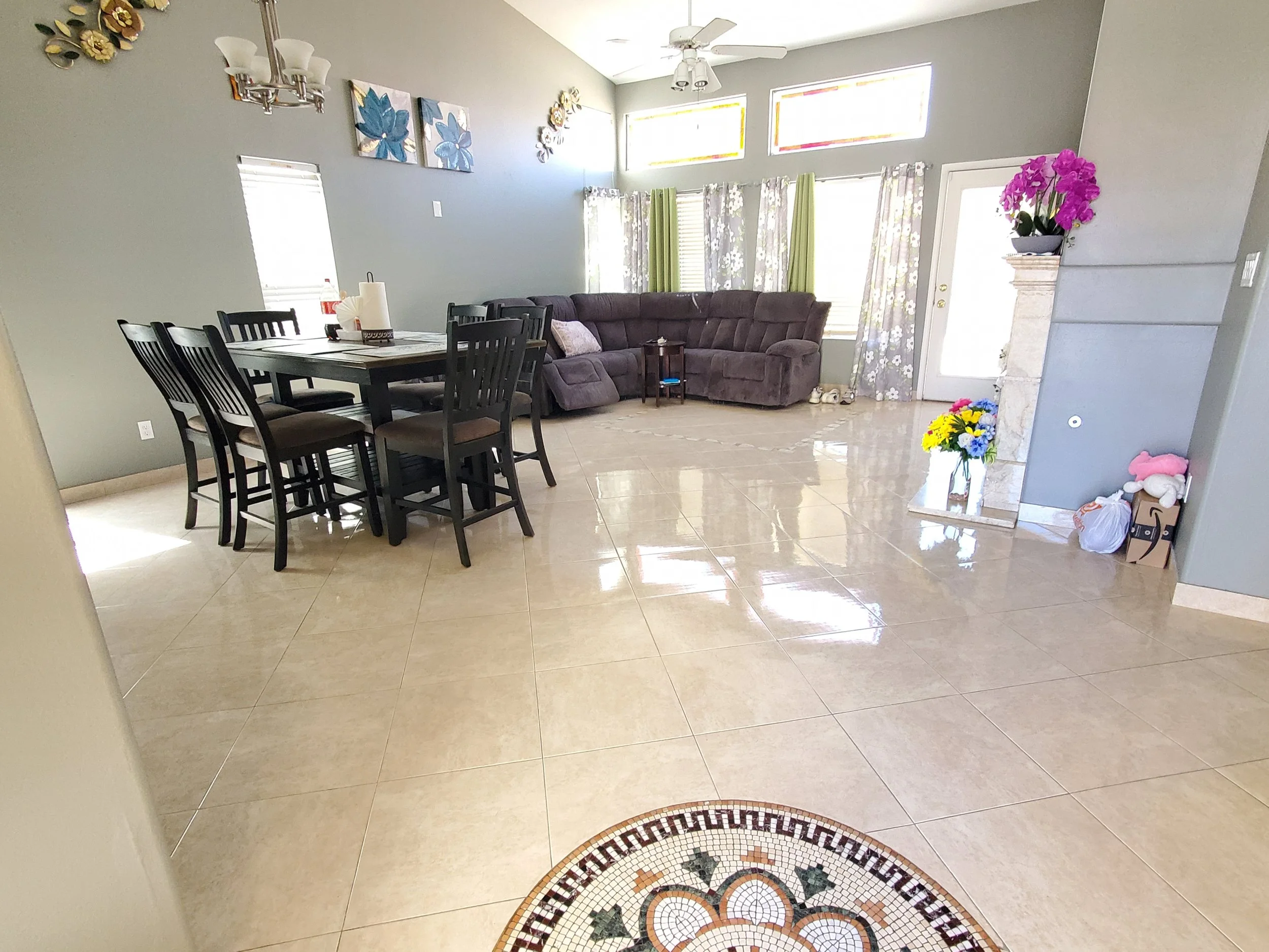 Living room with a dining table and chairs, a sectional sofa, and colorful curtains near large windows and a door, with floral wall art and decorative items.