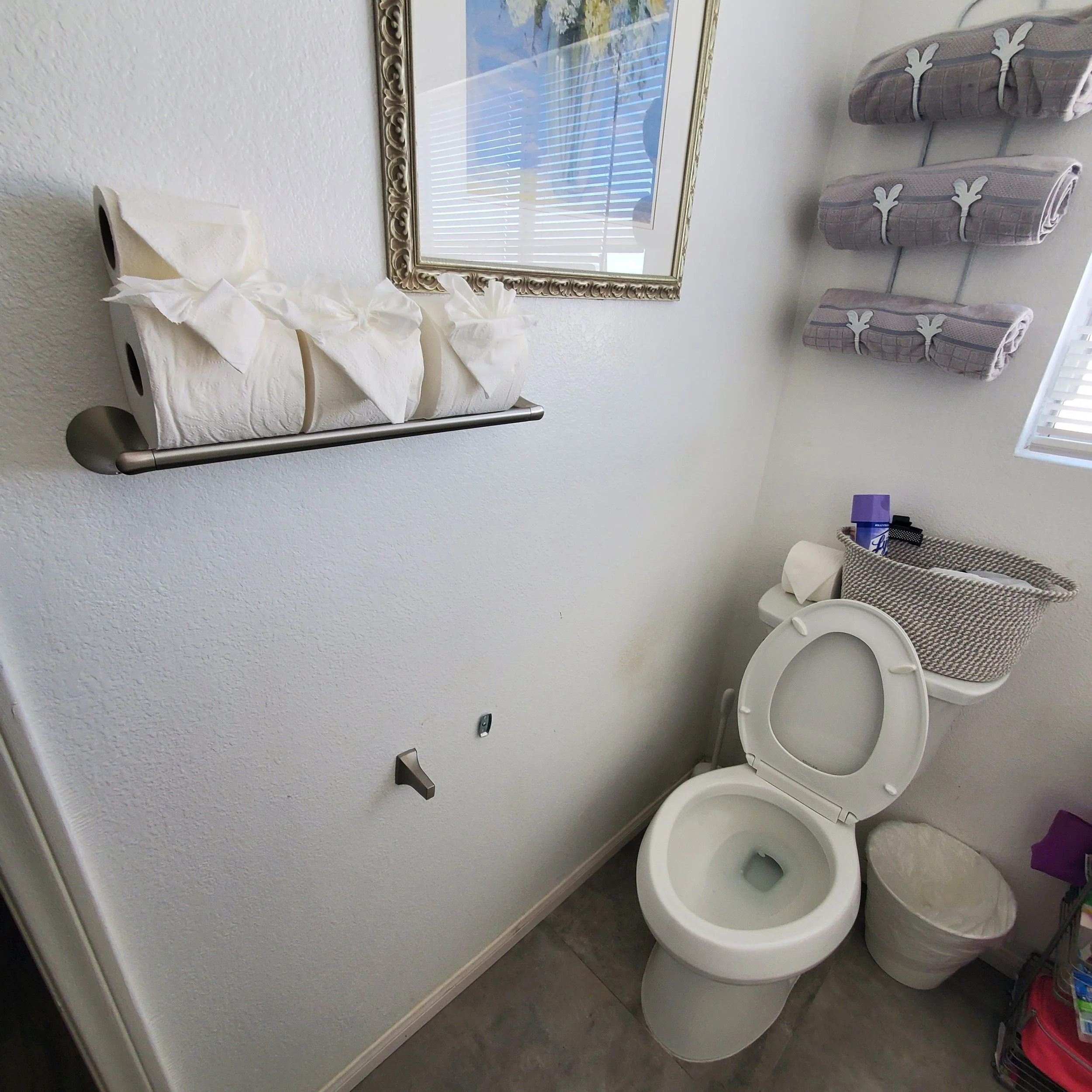 A public restroom with a white toilet, purple towel, and laundry baskets on the wall. There is a rectangular mirror with a decorative gold frame and a window with blinds above the toilet. The wall is painted white, and the floor is gray.