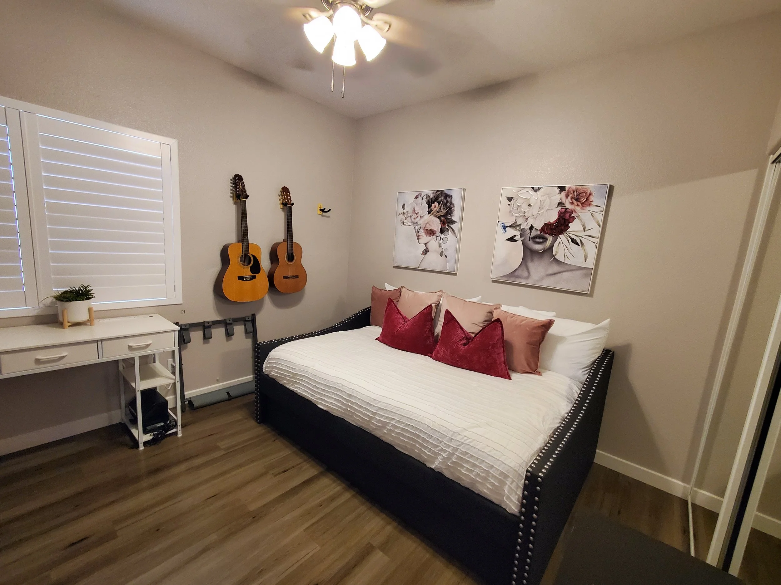 A bedroom with a bed, pink and burgundy pillows, two framed floral art pieces, two acoustic guitars hanging on the wall, a white desk with a plant, and a window with white shutters.