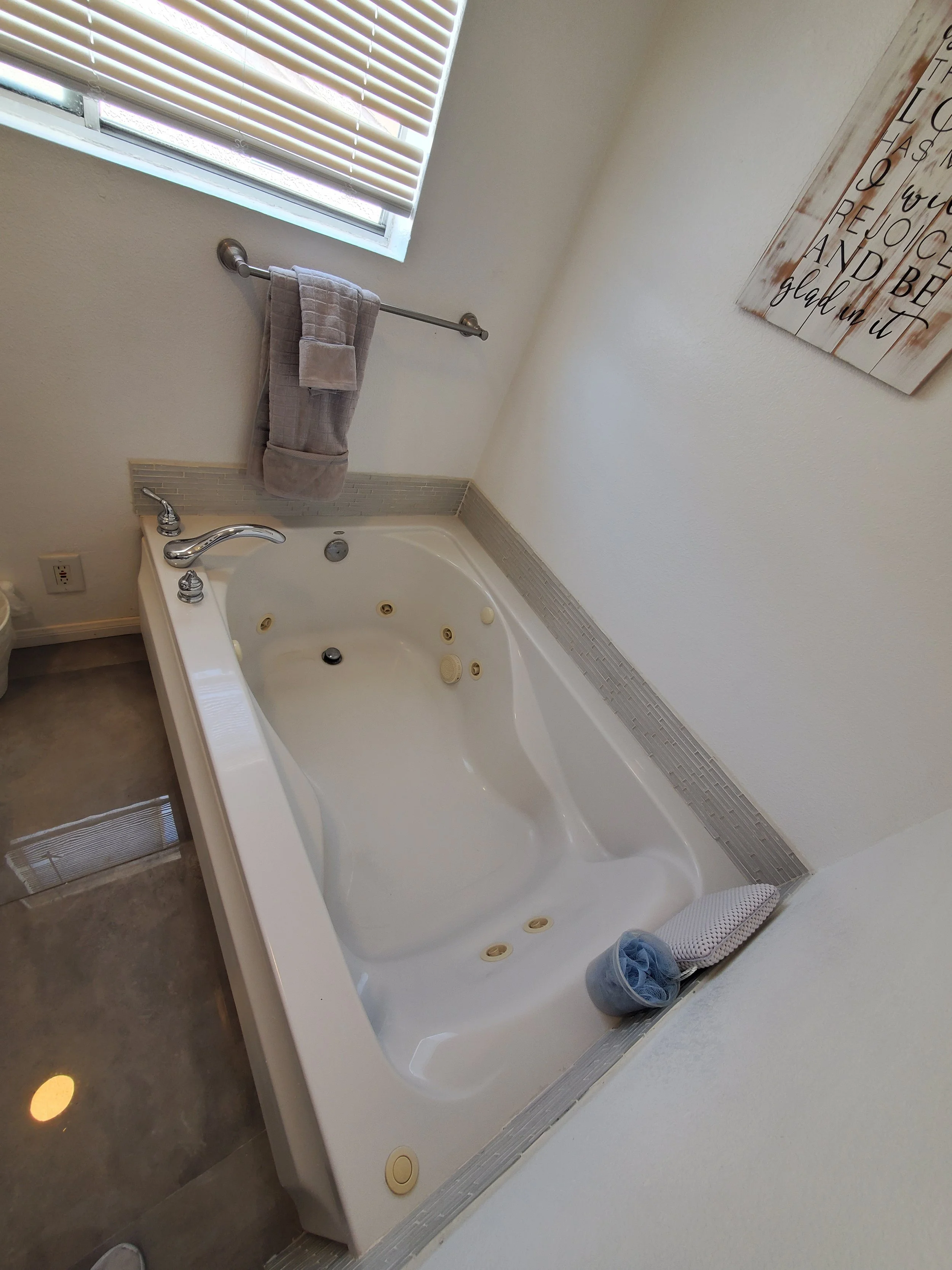 A bathroom with a white jacuzzi tub, a window with blinds, a gray towel hanging on a metal rail, a sign on the wall with an inspirational message, and a rolled-up towel on the floor near the tub.