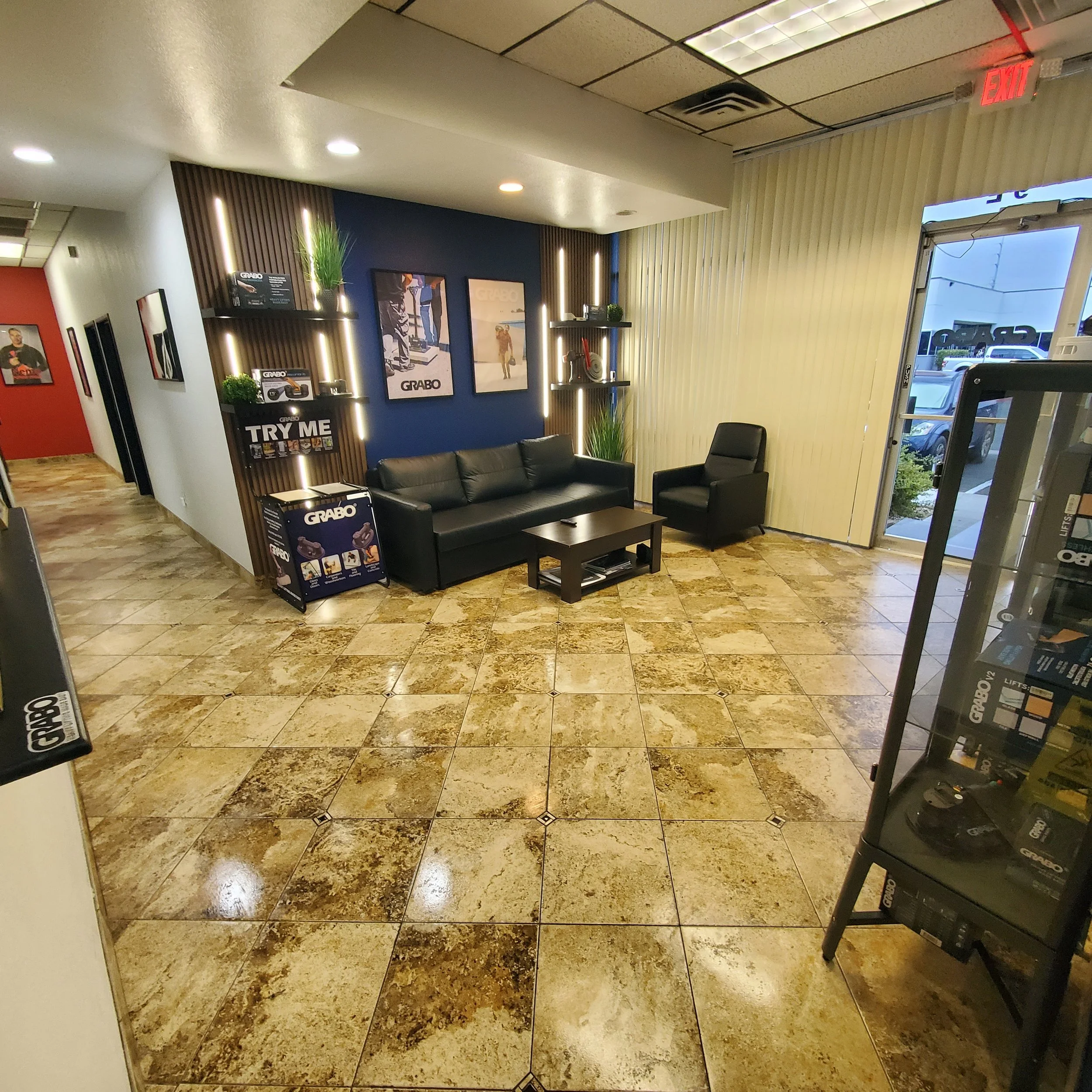 A waiting area inside a commercial building with tan marble flooring, black leather couches, tall green plants, and framed posters on the wall. Part of the entrance door is visible on the right, with an illuminated red exit sign above it.