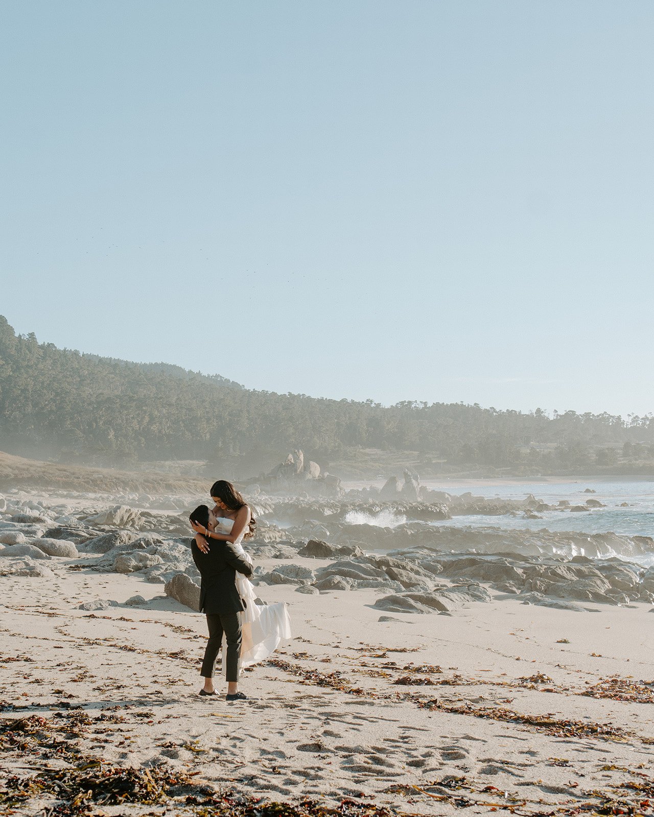 A couple on a beach, with the man lifting the woman in front of rocky shoreline and ocean waves, amidst a scenic landscape with hills covered in trees.