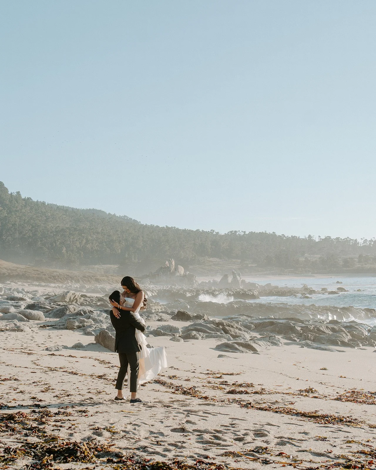 Groom lifting his bride into the air in the sand. with the beach coast line in the background.