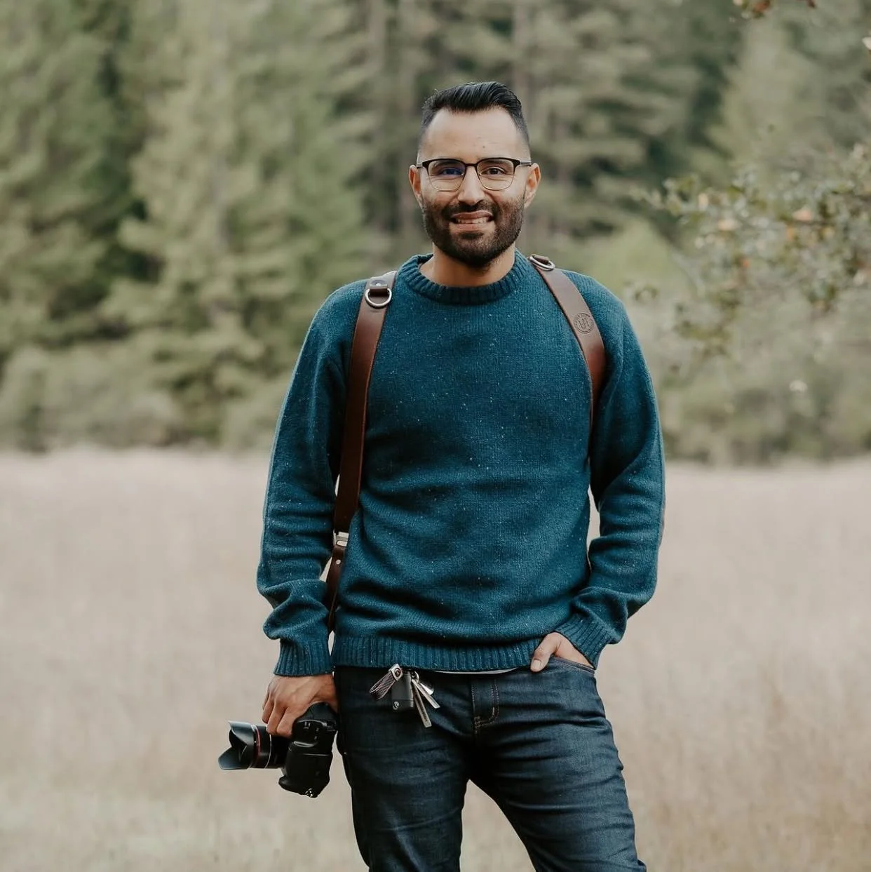 A man with glasses, a beard, and short black hair, standing outdoors with trees in the background. He is wearing a blue sweater, black pants, and has a camera hanging from his right hand. He also has a brown backpack over his shoulders, and his left hand is in his pocket.