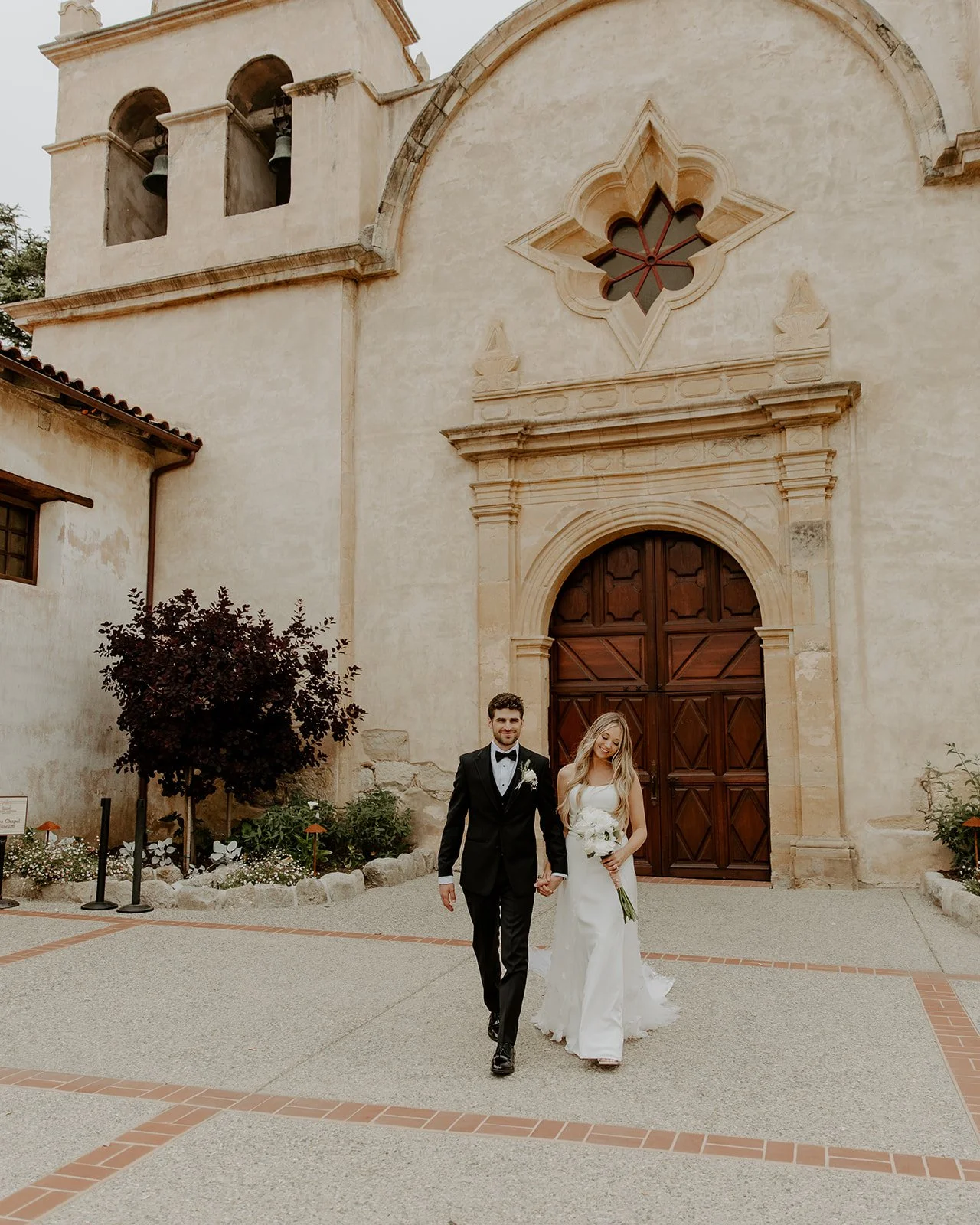 A bride and groom walking hand in hand outside a historic church, with the bride holding a bouquet and both dressed in wedding attire.