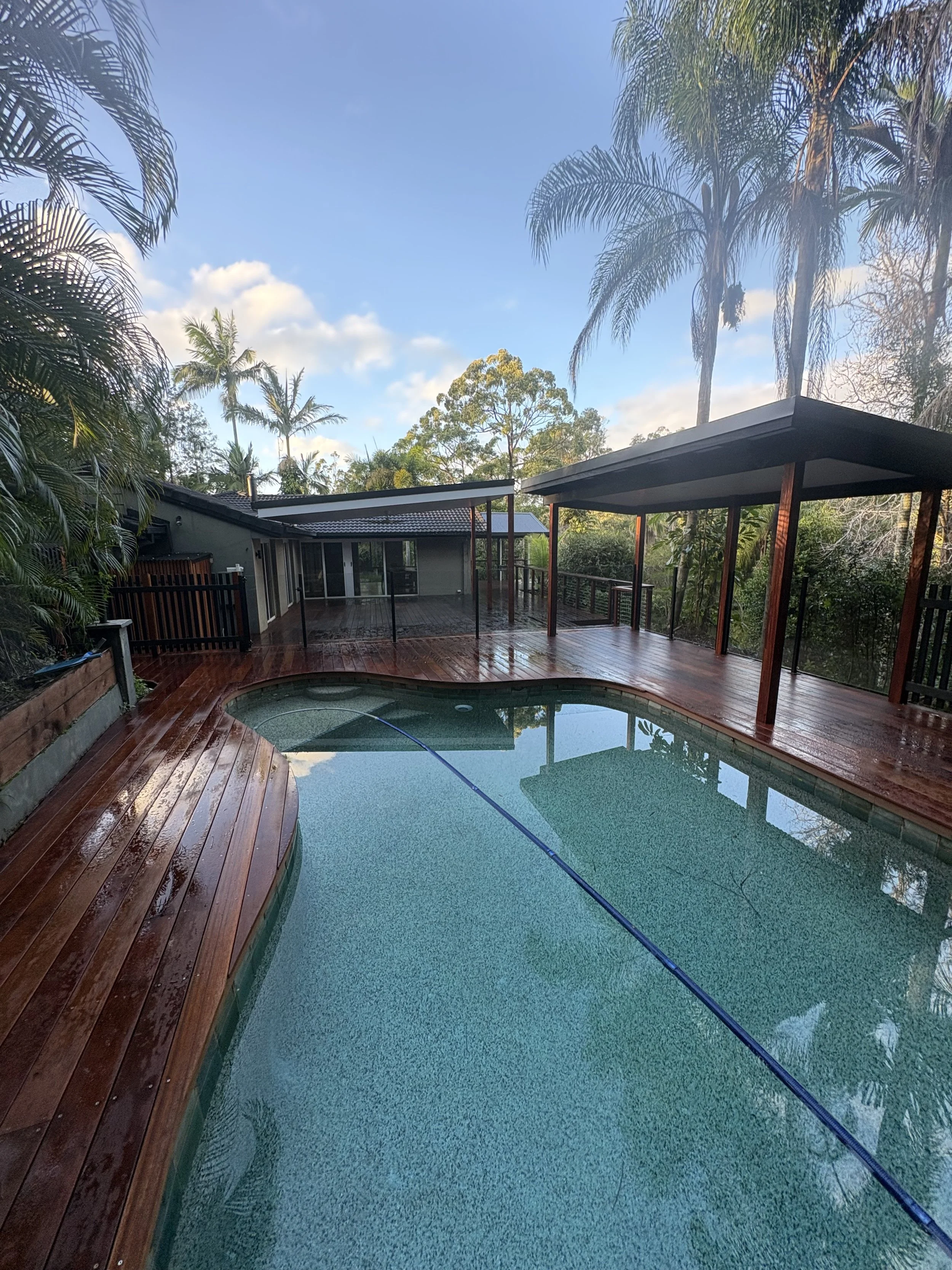 An outdoor scene with a house, a swimming pool, and a deck surrounded by lush green trees and palm trees under a partly cloudy sky.